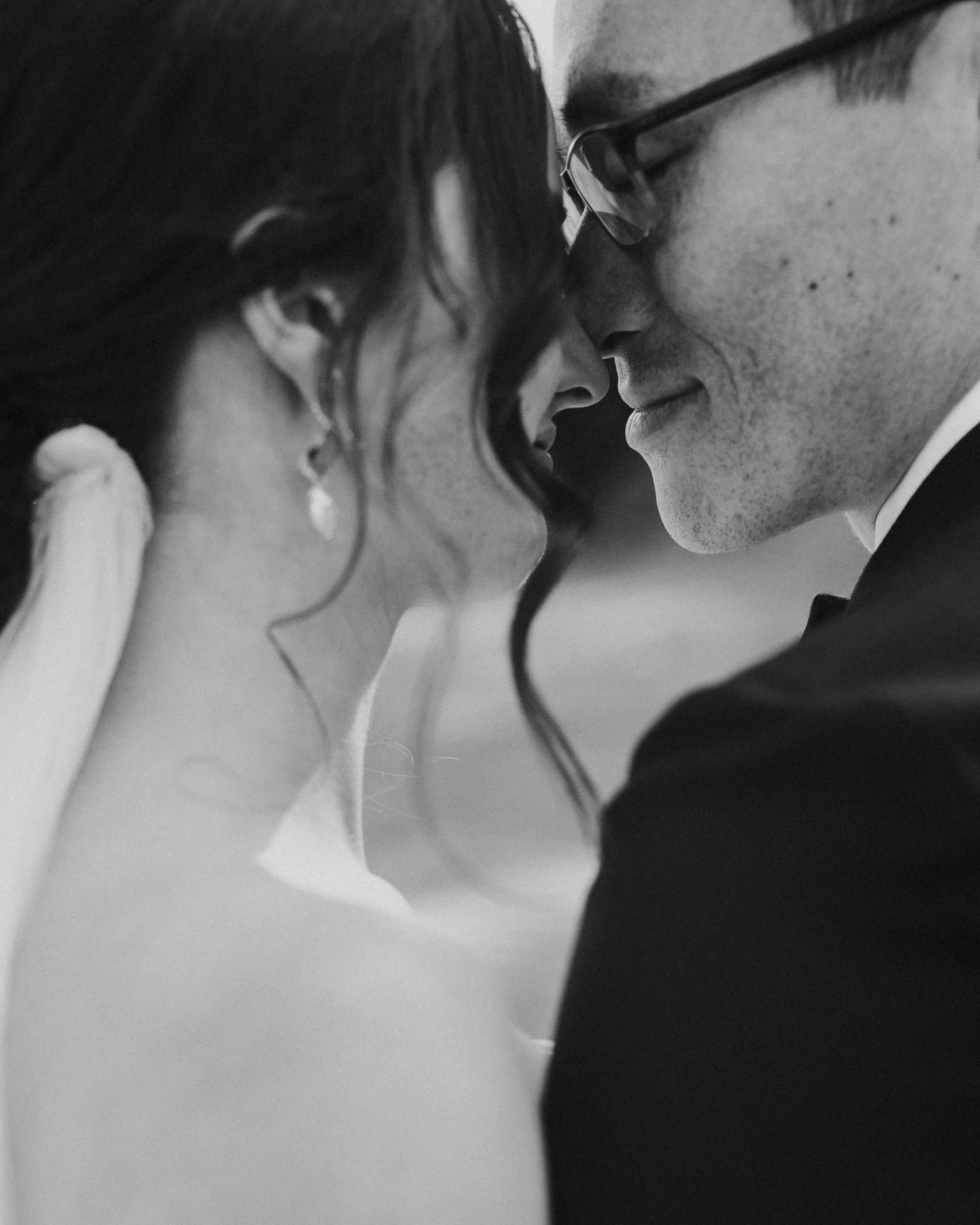 A close-up black and white photo of a couple with their faces touching, showing an intimate moment, likely at a wedding or special event.