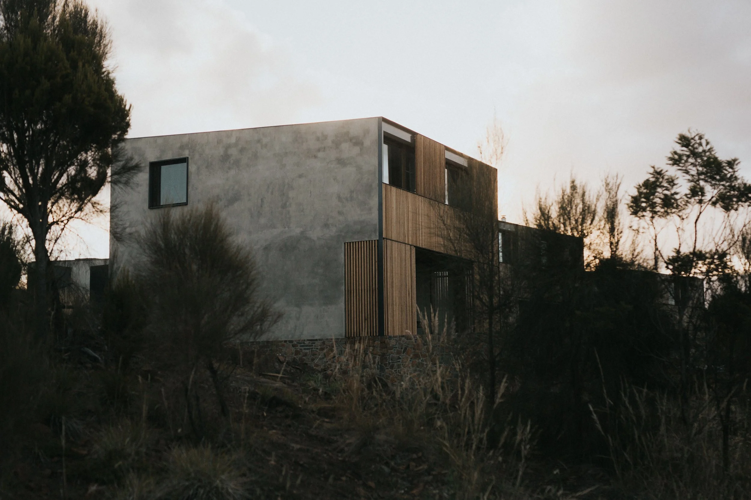 Modern two-story house with minimalist design, situated among trees and bushes, with a stone foundation and wooden accents, at dusk.