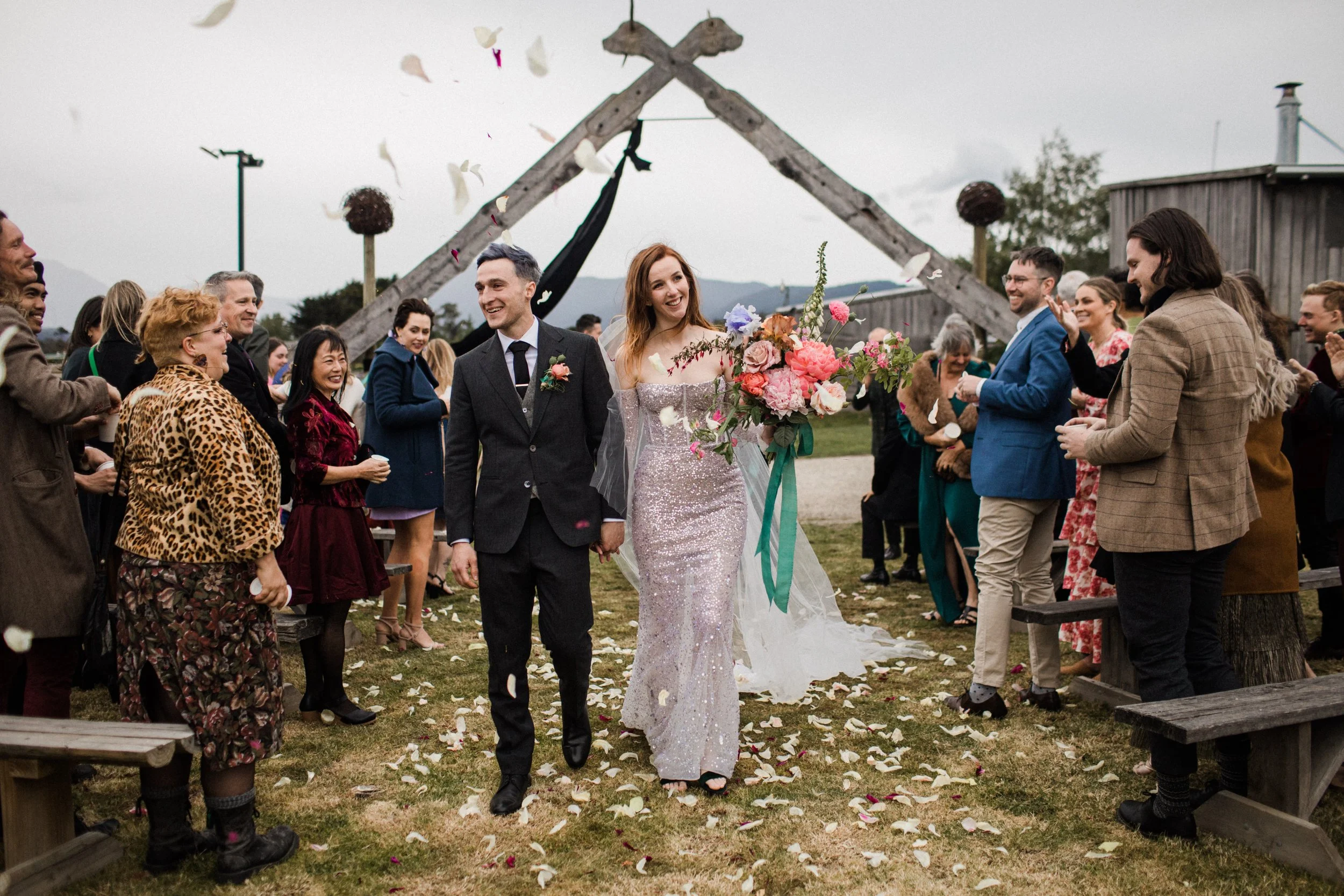 A bride and groom walking hand in hand down the aisle at an outdoor wedding, surrounded by smiling guests, with flower petals on the ground and a rustic wooden arch in the background.