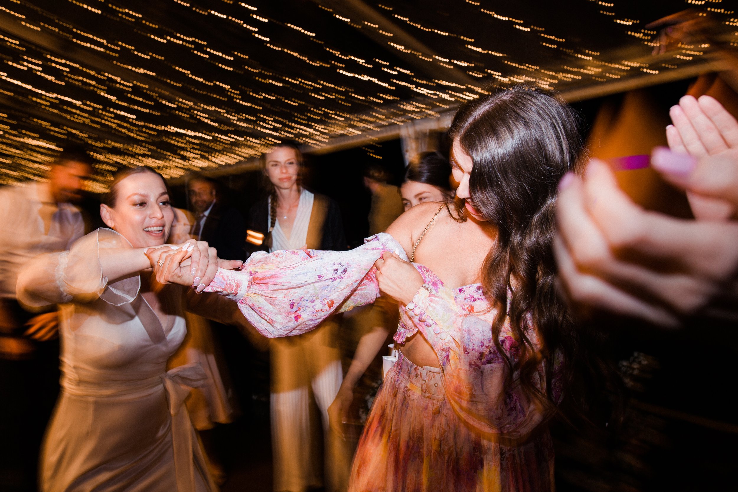 People dancing and holding hands at a lively indoor event with string lights on the ceiling.