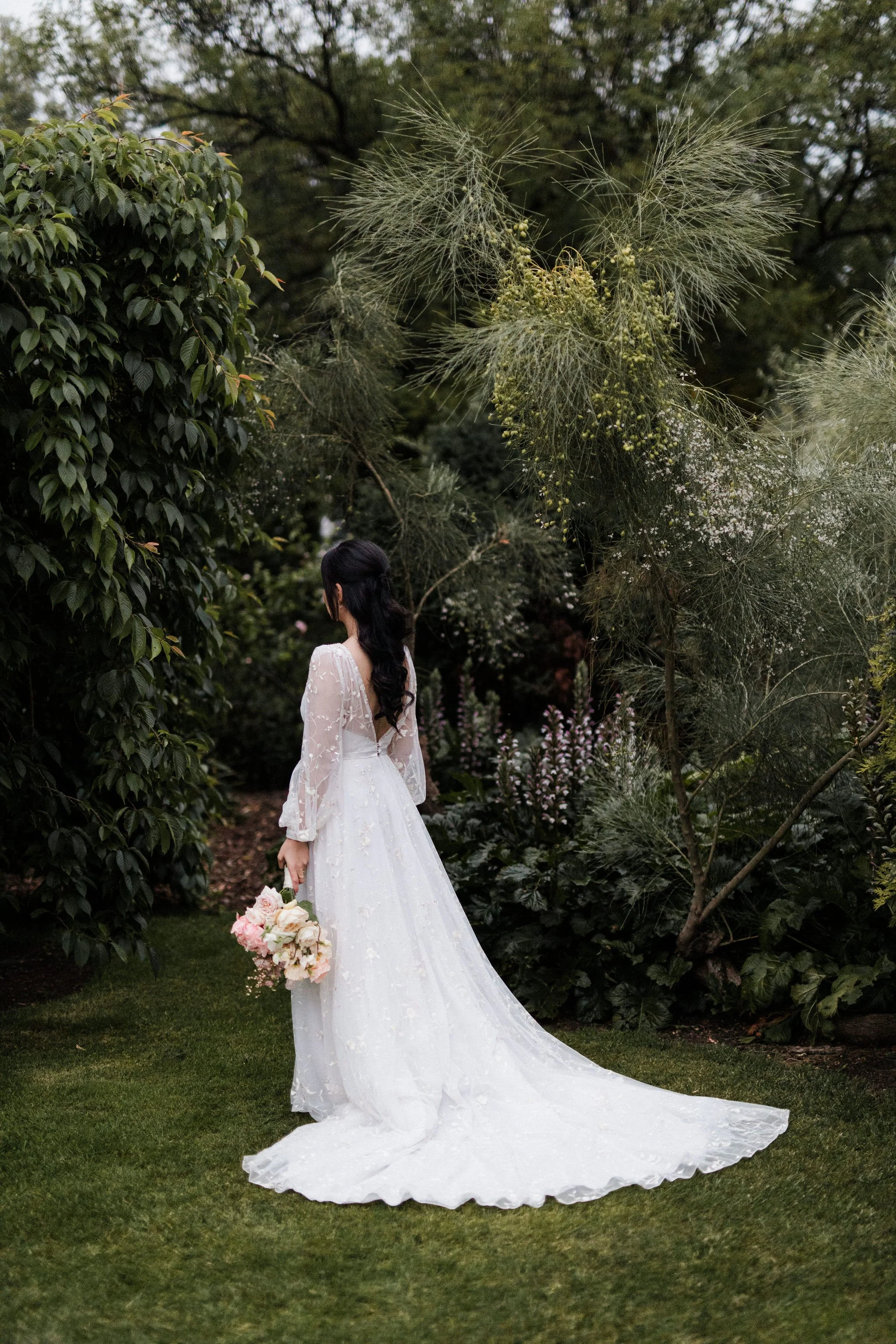 A bride in a white wedding dress holding a pink and white bouquet standing on grass in a garden surrounded by lush greenery and flowering plants.