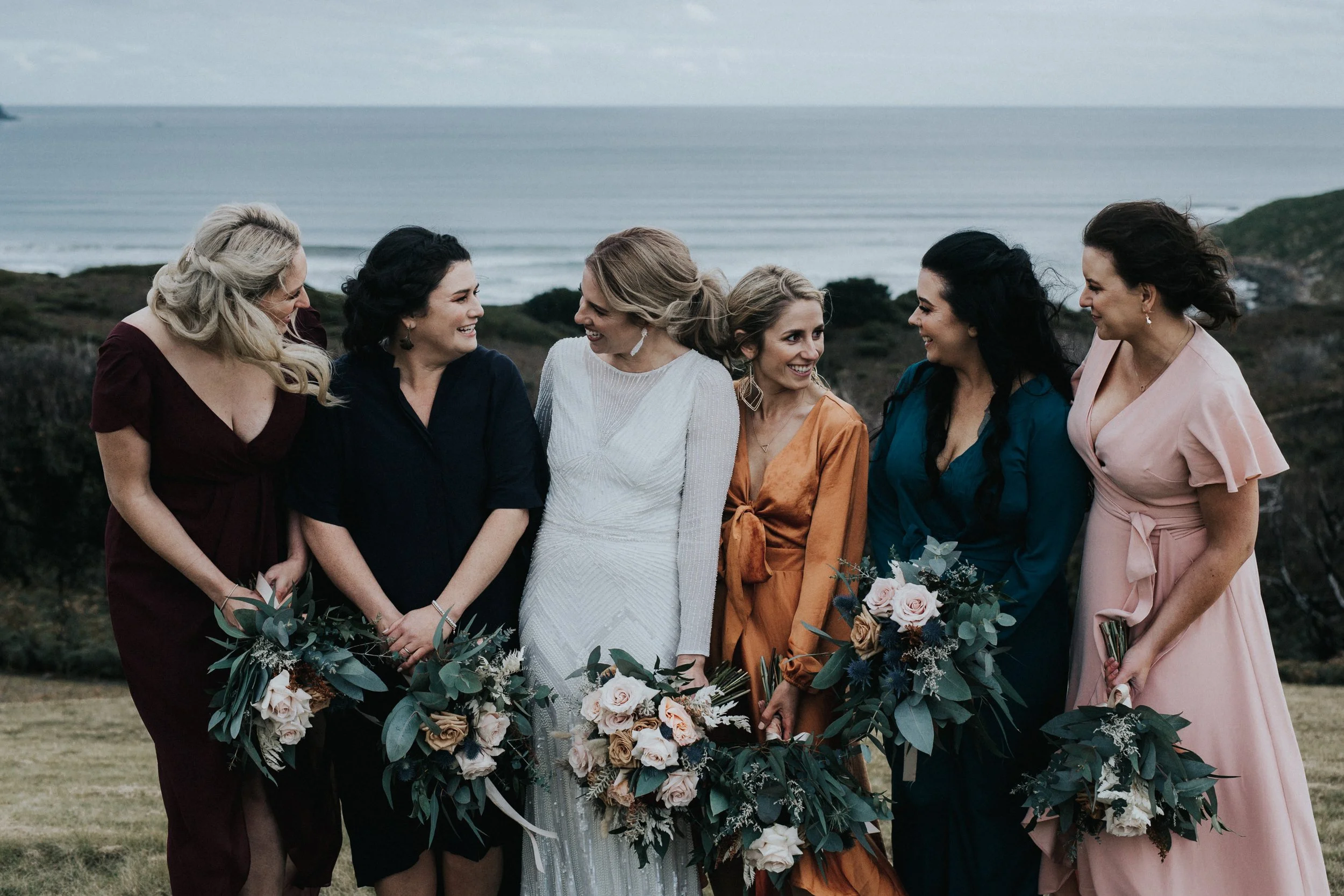 Group of seven women dressed in colorful dresses standing outdoors with ocean in the background, holding bouquets of flowers and smiling at each other.
