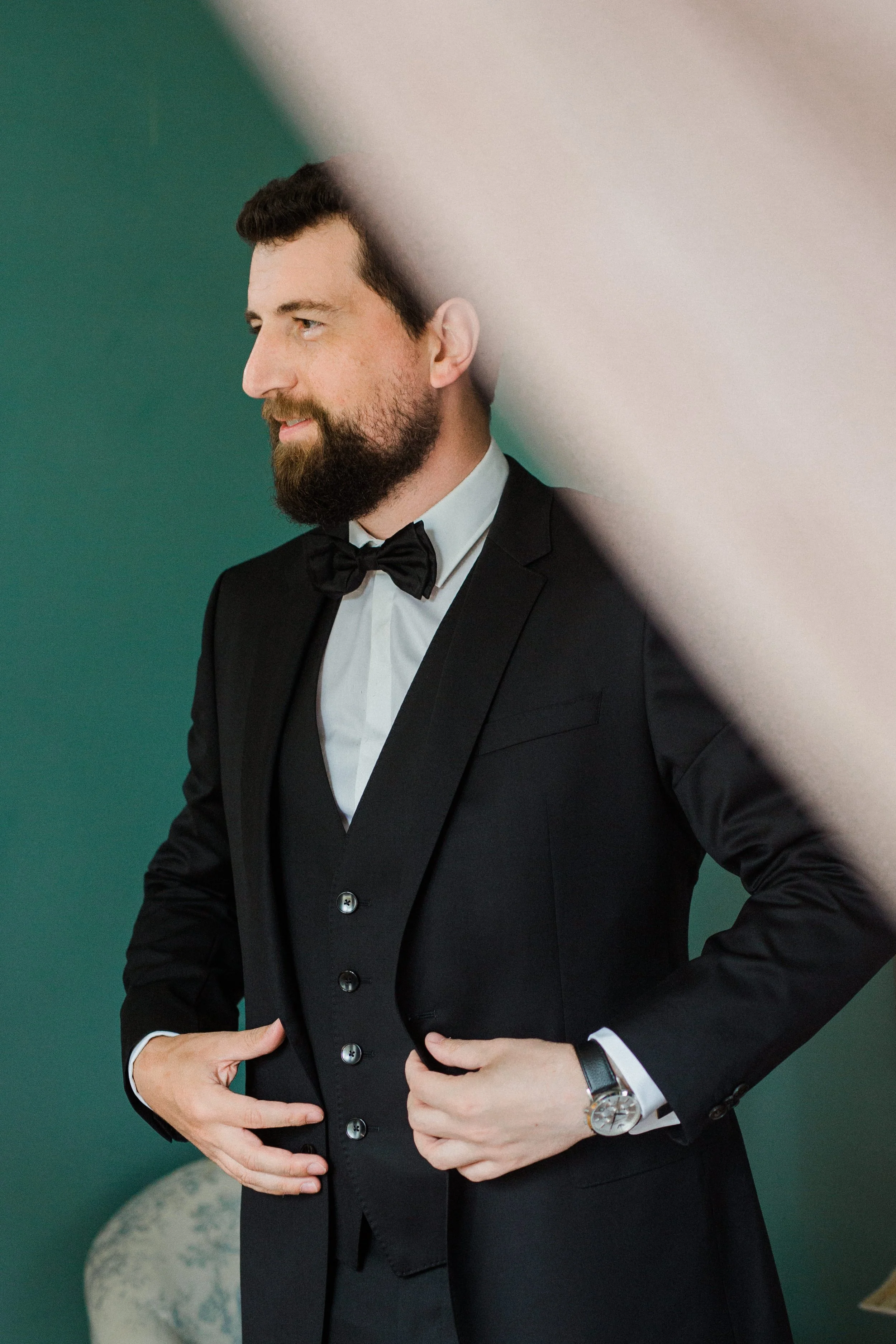 A man in a black tuxedo and bow tie adjusting his vest, standing against a teal wall with a partially visible beige decorative object in the background.