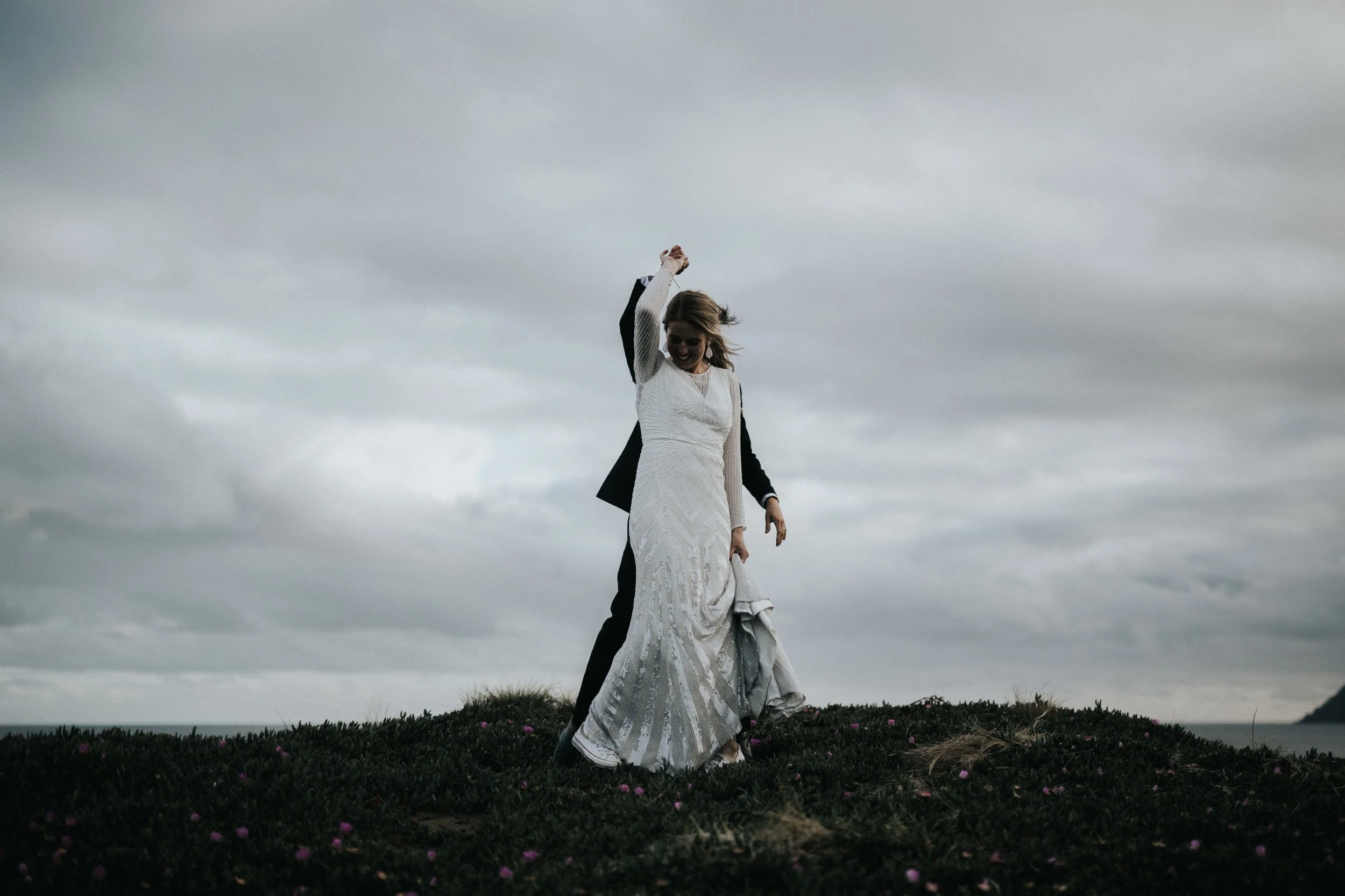 A woman in a wedding dress holding up a man dressed in a suit behind her, standing outdoors on a grassy hill with cloudy sky in the background.