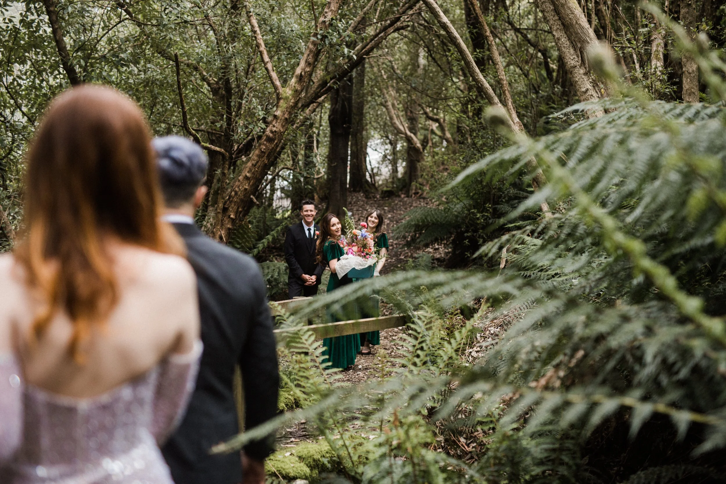 A wedding ceremony taking place outdoors in a forest, with a couple in the foreground and the officiant and two bridesmaids holding a bouquet in the background.