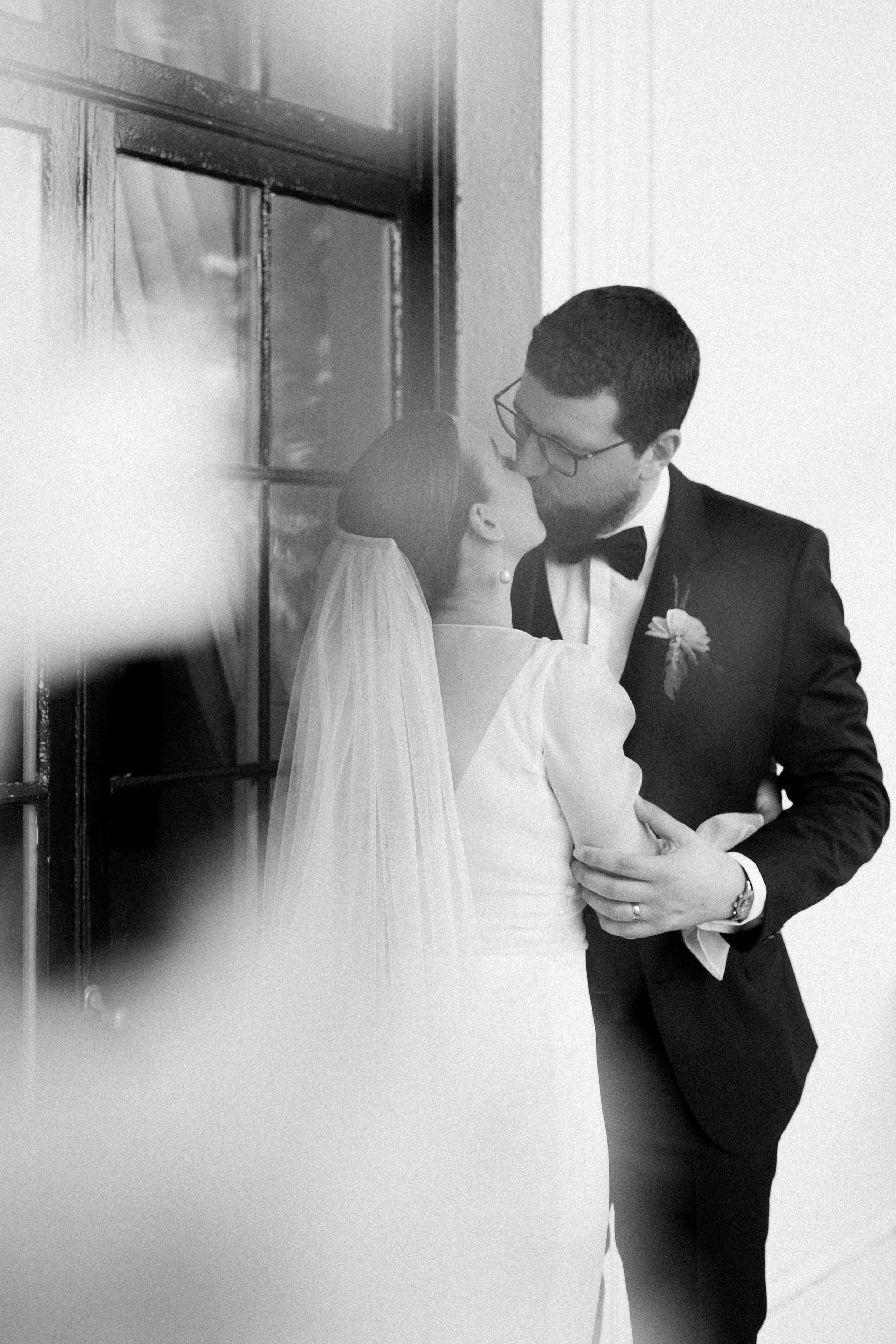 A black-and-white photo of a bride and groom sharing a kiss during their wedding, with the groom embracing the bride.