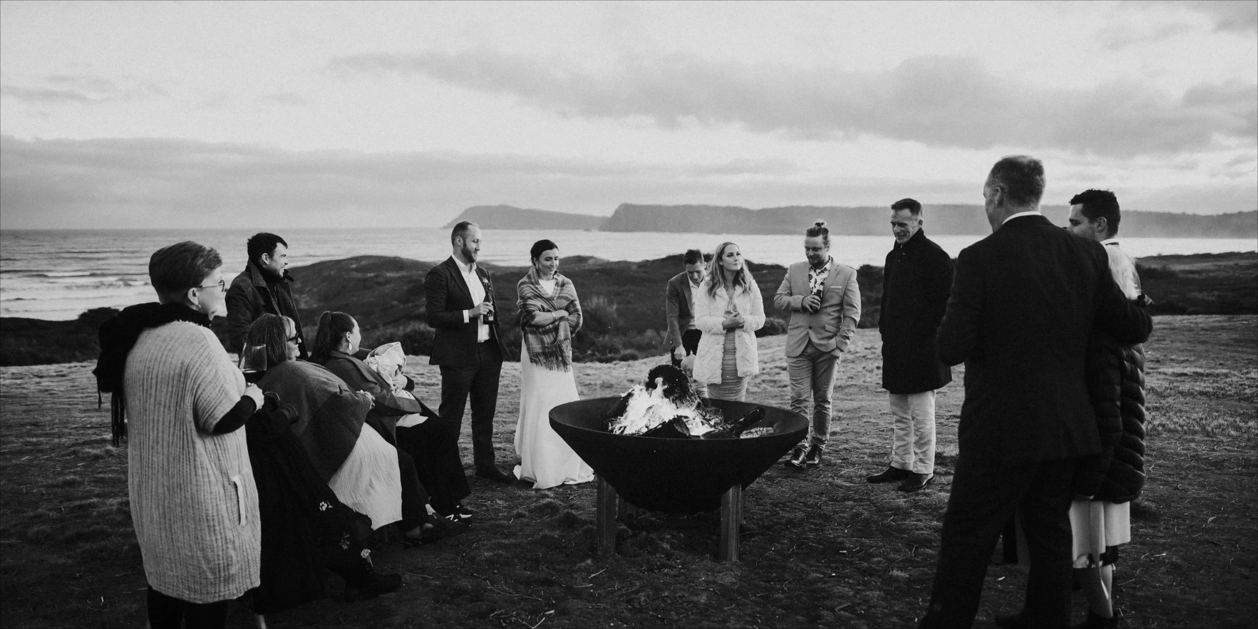 People gathered around a fire on a beach, possibly for a social event or gathering, with the ocean and cliffs in the background, in black and white.