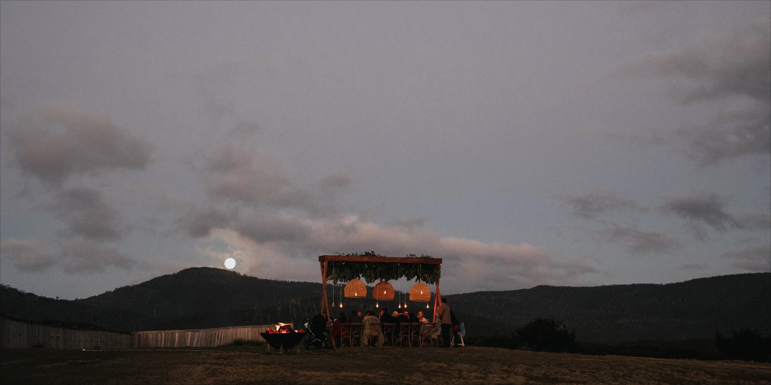 People gathered around a table under a wooden canopy with hanging lanterns during dusk, with mountains and a partly cloudy sky and a bright full moon in the background.