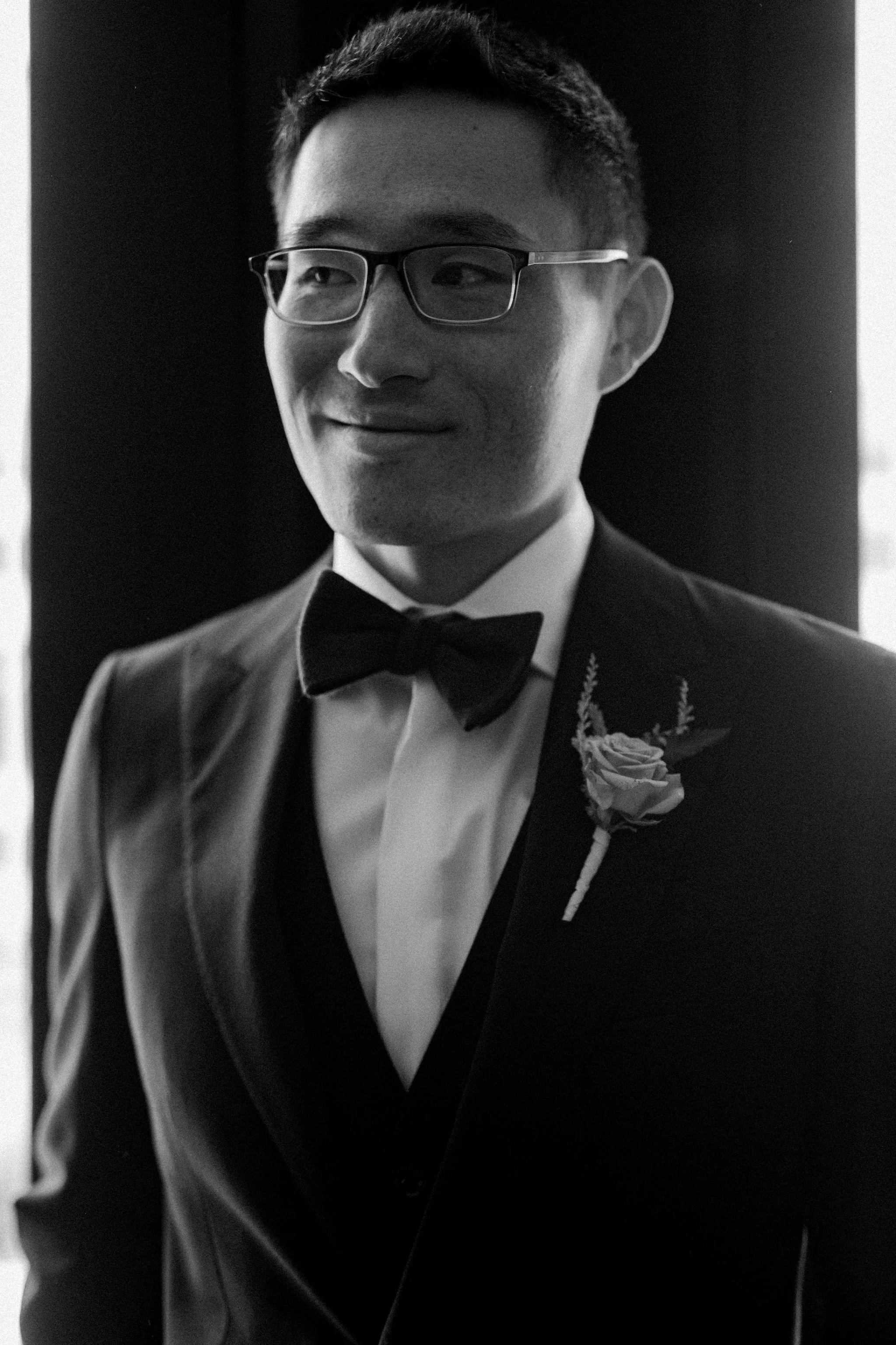 A man wearing glasses, a tuxedo with a bowtie, and a boutonniere, smiling slightly in a black-and-white photo.
