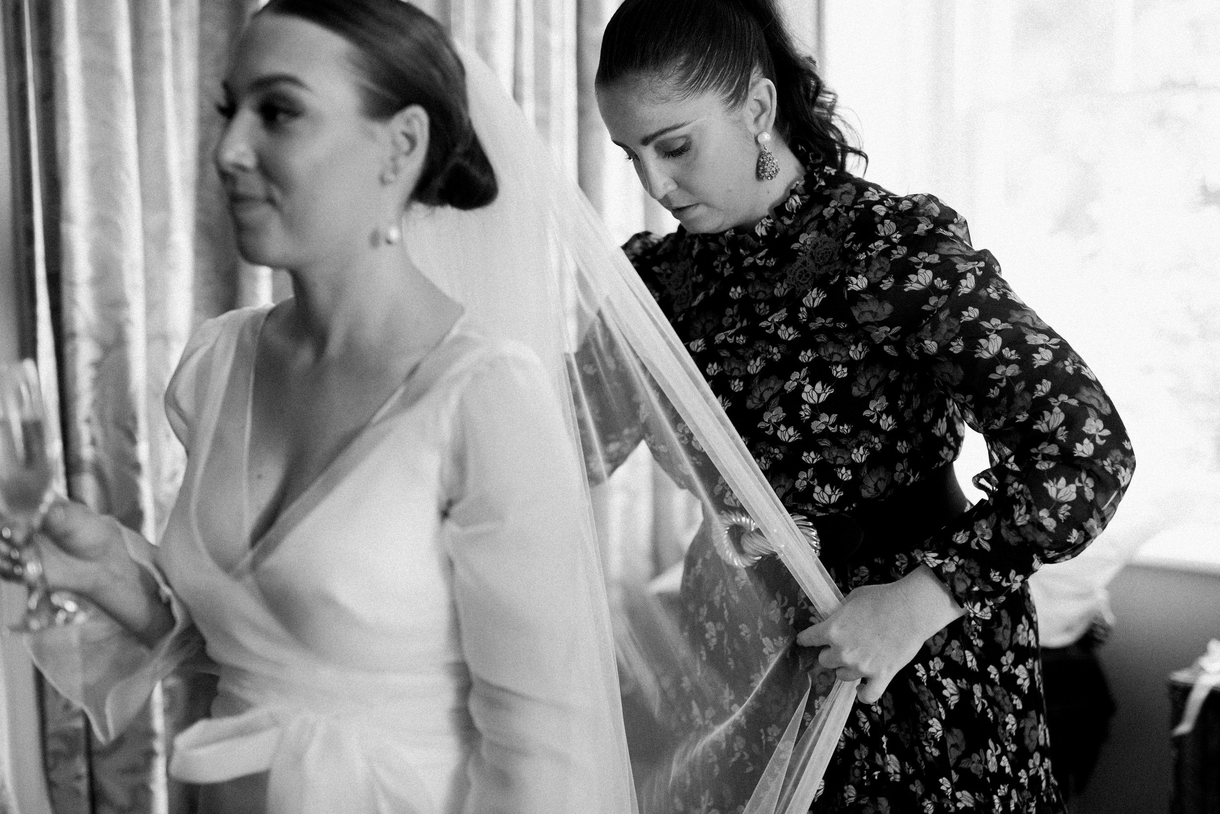 A woman in a white dress with a veil is getting ready for her wedding, assisted by another woman in a black patterned dress who is holding a frame or picture.