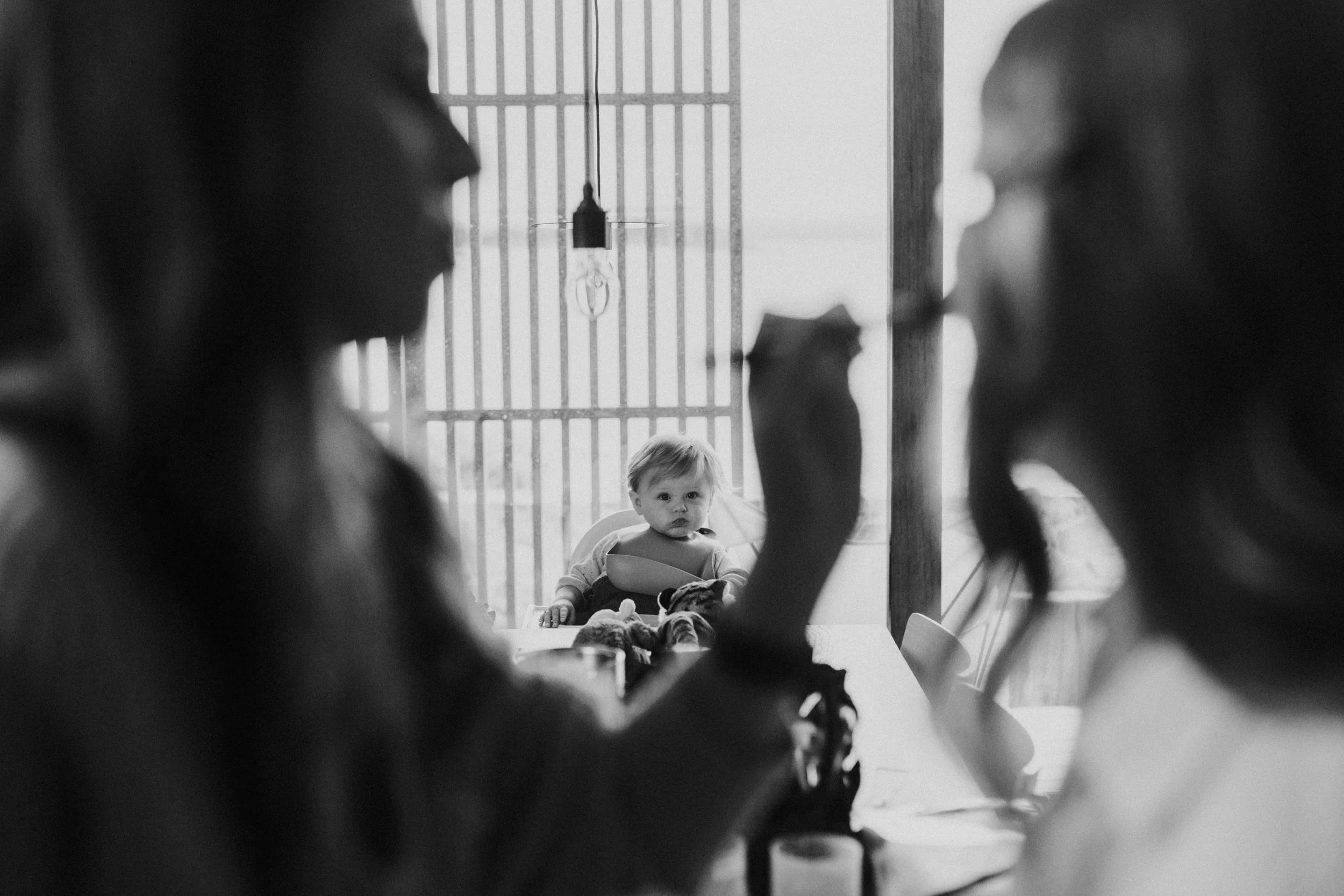 Two women are sitting at a table, engaged in a conversation, with a young child seated in relief in the background looking toward the camera.