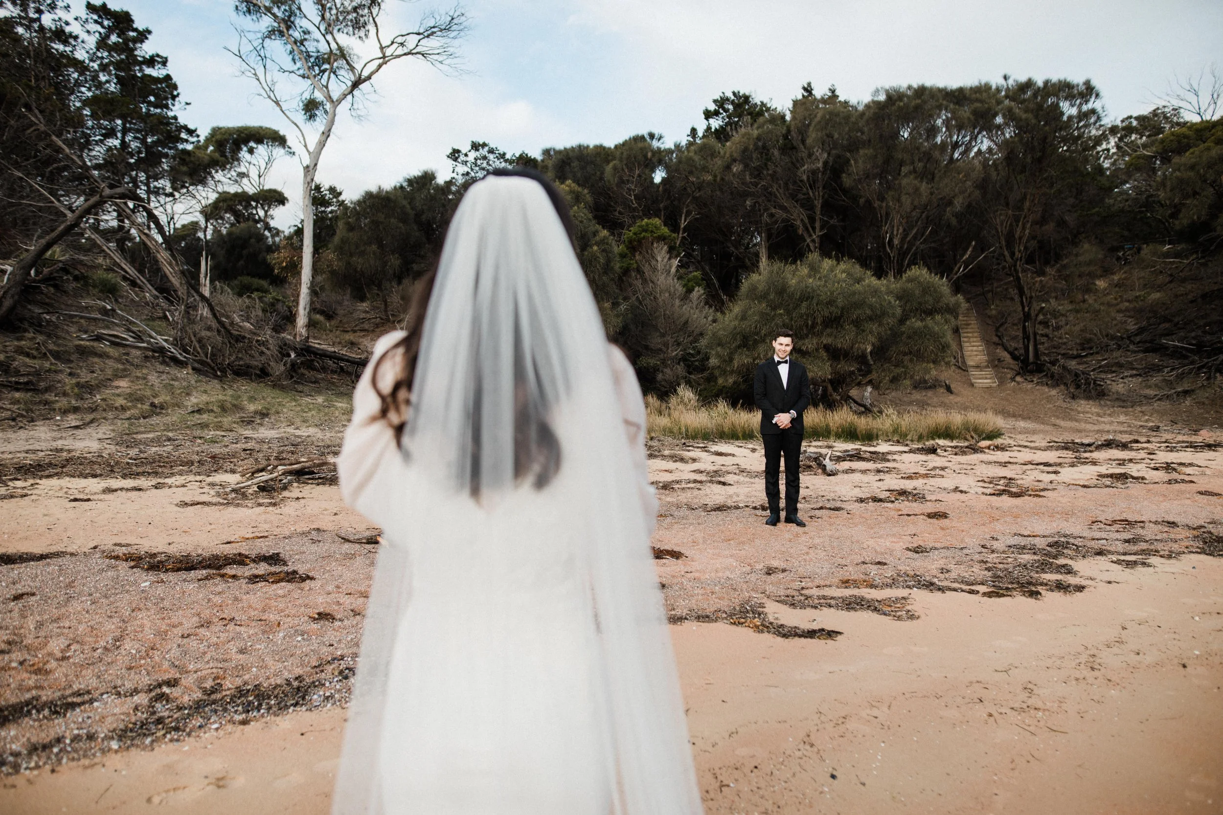 A bride with a veil stands on the beach facing a groom in a tuxedo, with trees and a staircase in the background.