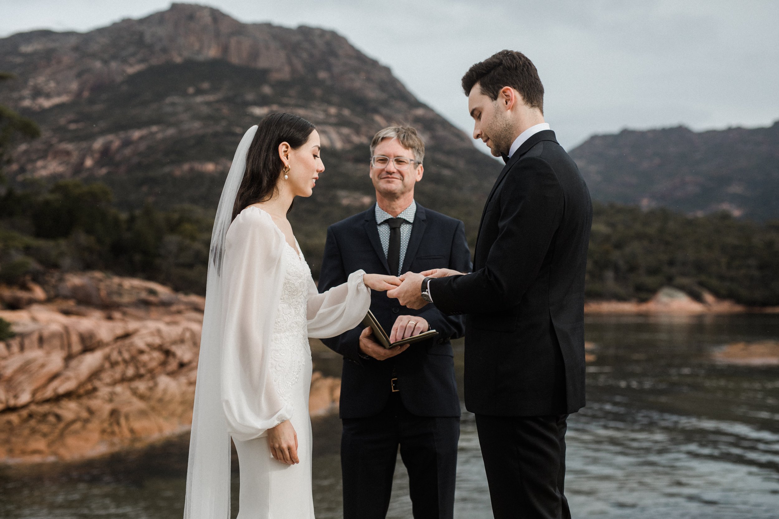 A couple gets married outdoors in front of a mountain and water, with an officiant holding a book, as they exchange rings and hold hands.