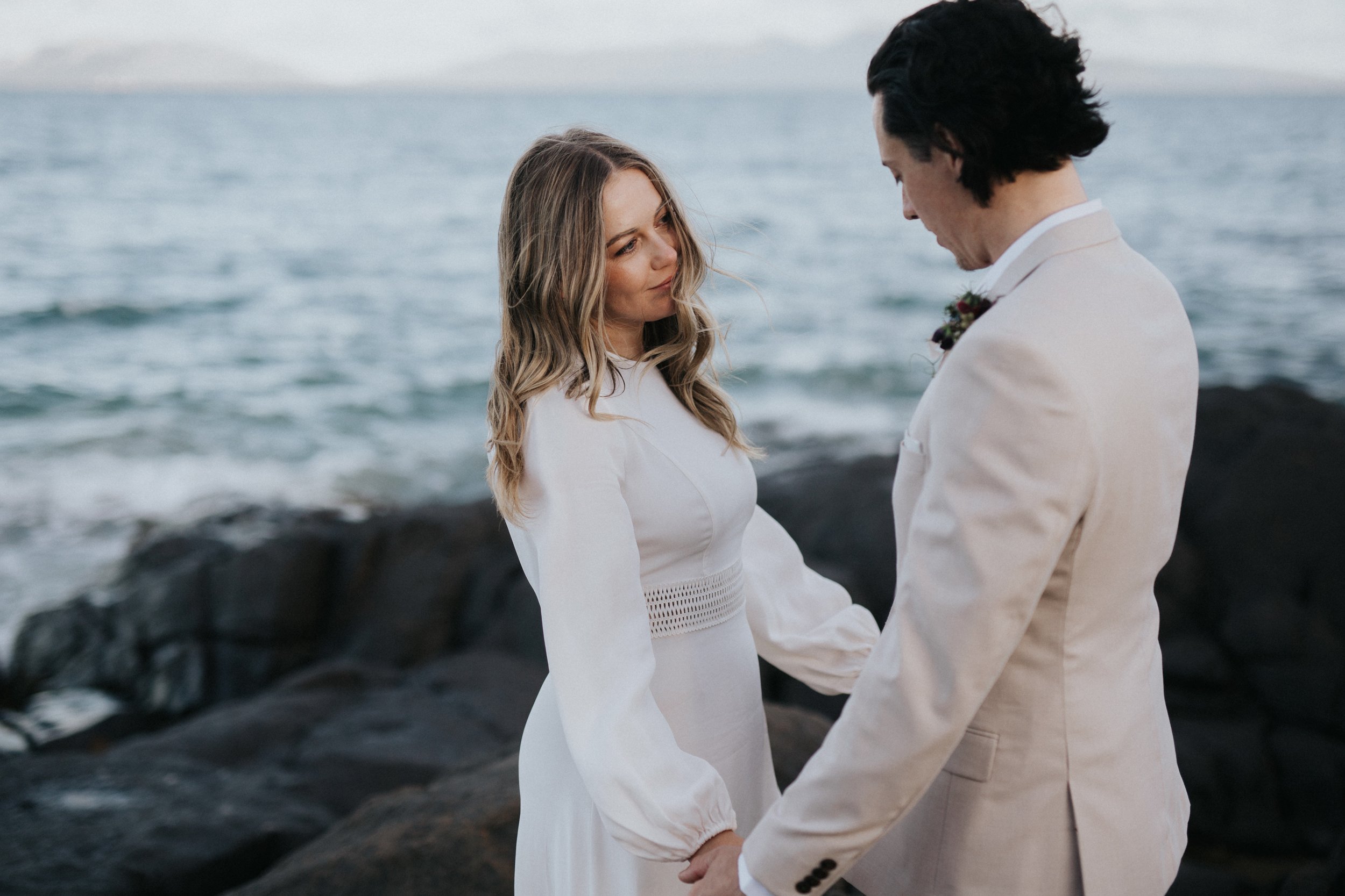 A couple in wedding attire holding hands on a rocky shoreline, with the ocean in the background.