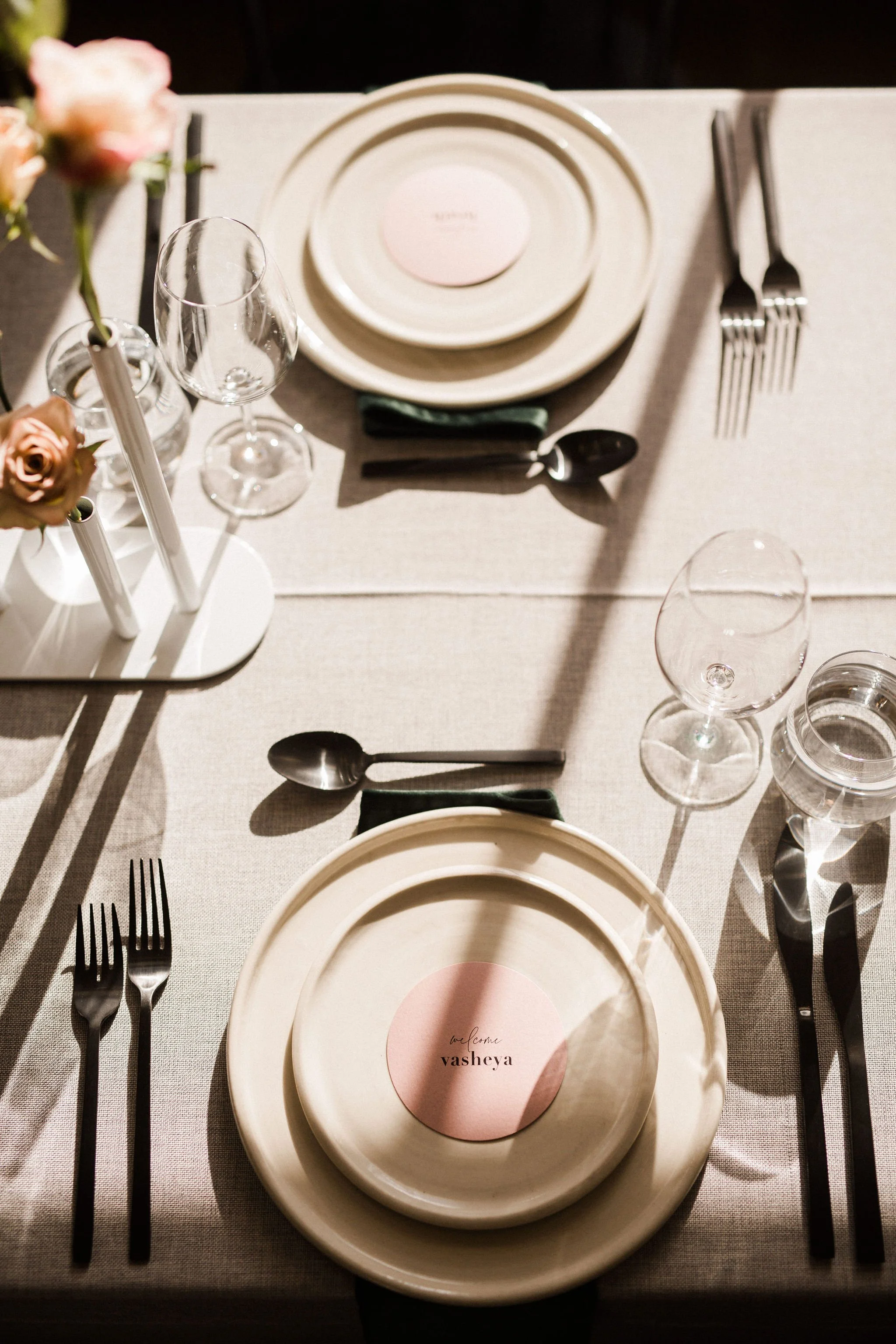 Elegant table setting with beige plates, black cutlery, wine glasses, water glasses, and a note that reads 'love from vasheya' on pink and white circular paper, on a light-colored tablecloth.