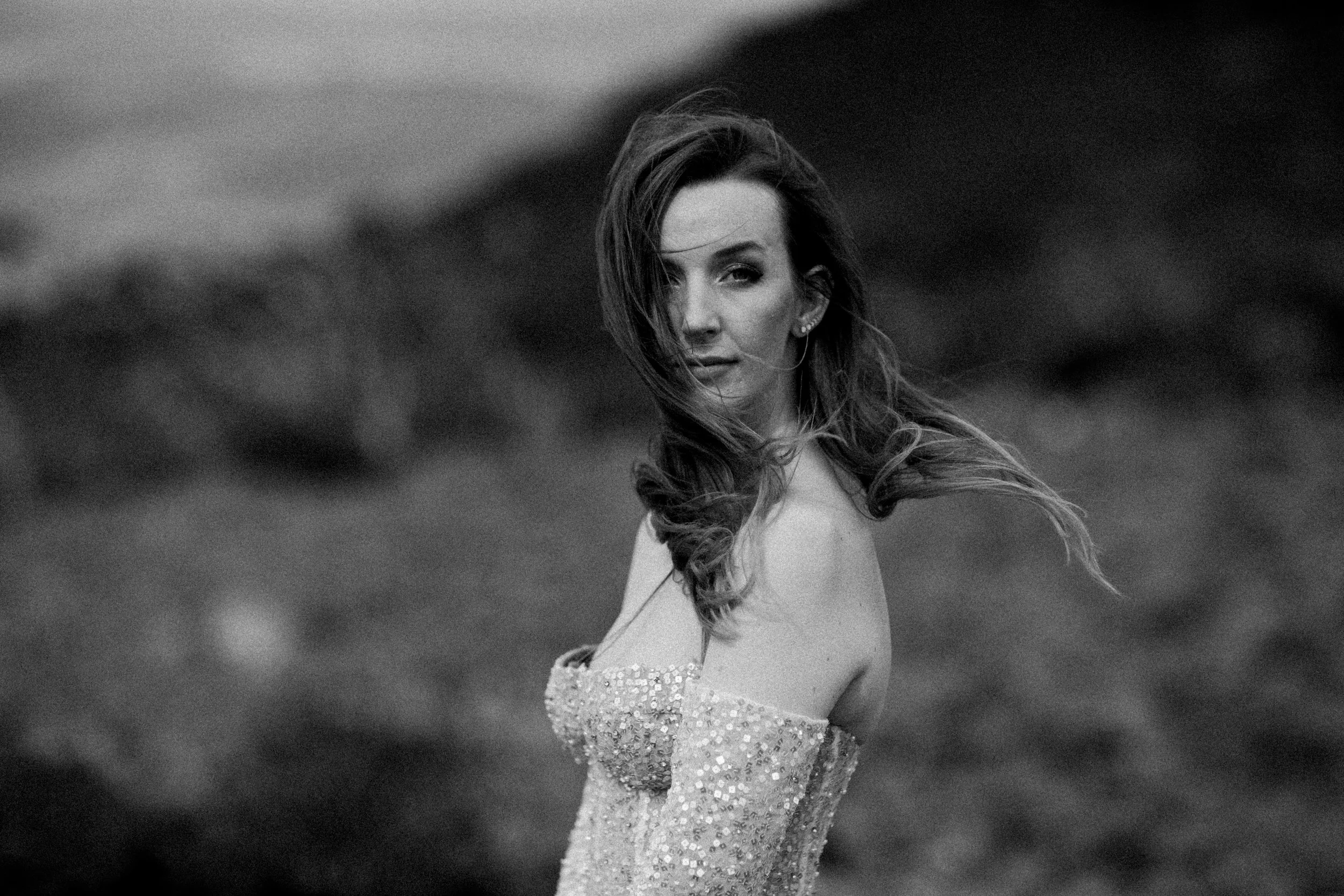 Black and white photo of a woman with long hair, wearing an off-the-shoulder dress with sequins, standing outdoors with a blurred natural background.
