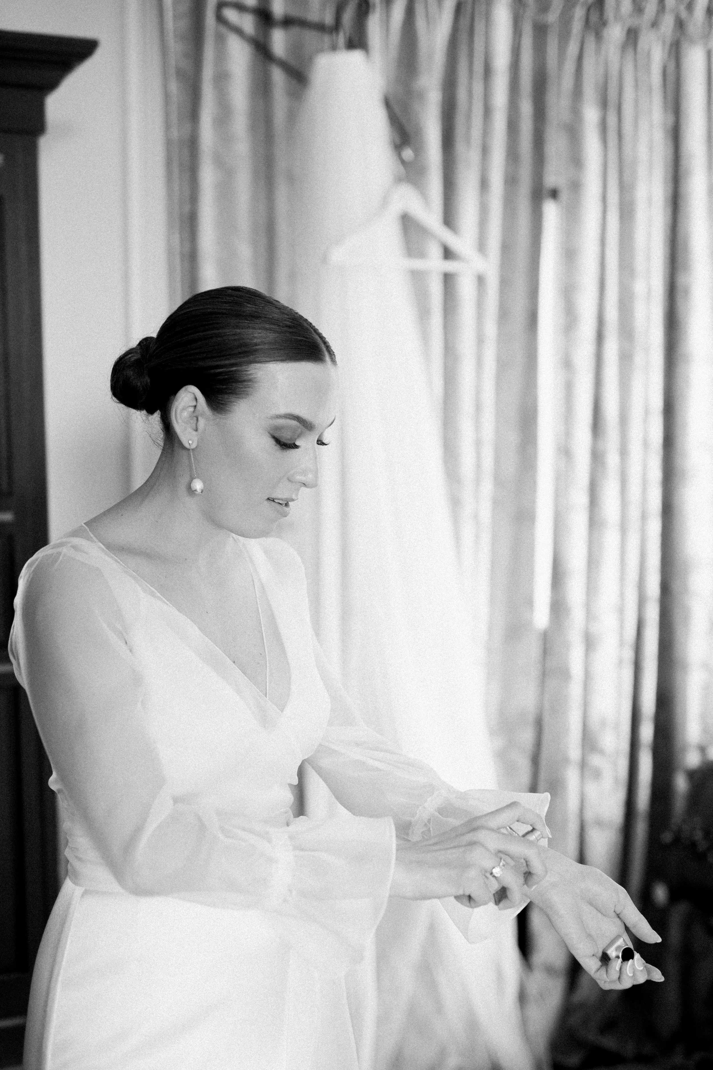 A woman in a wedding dress preparing in a room, with a wedding gown hanging behind her.