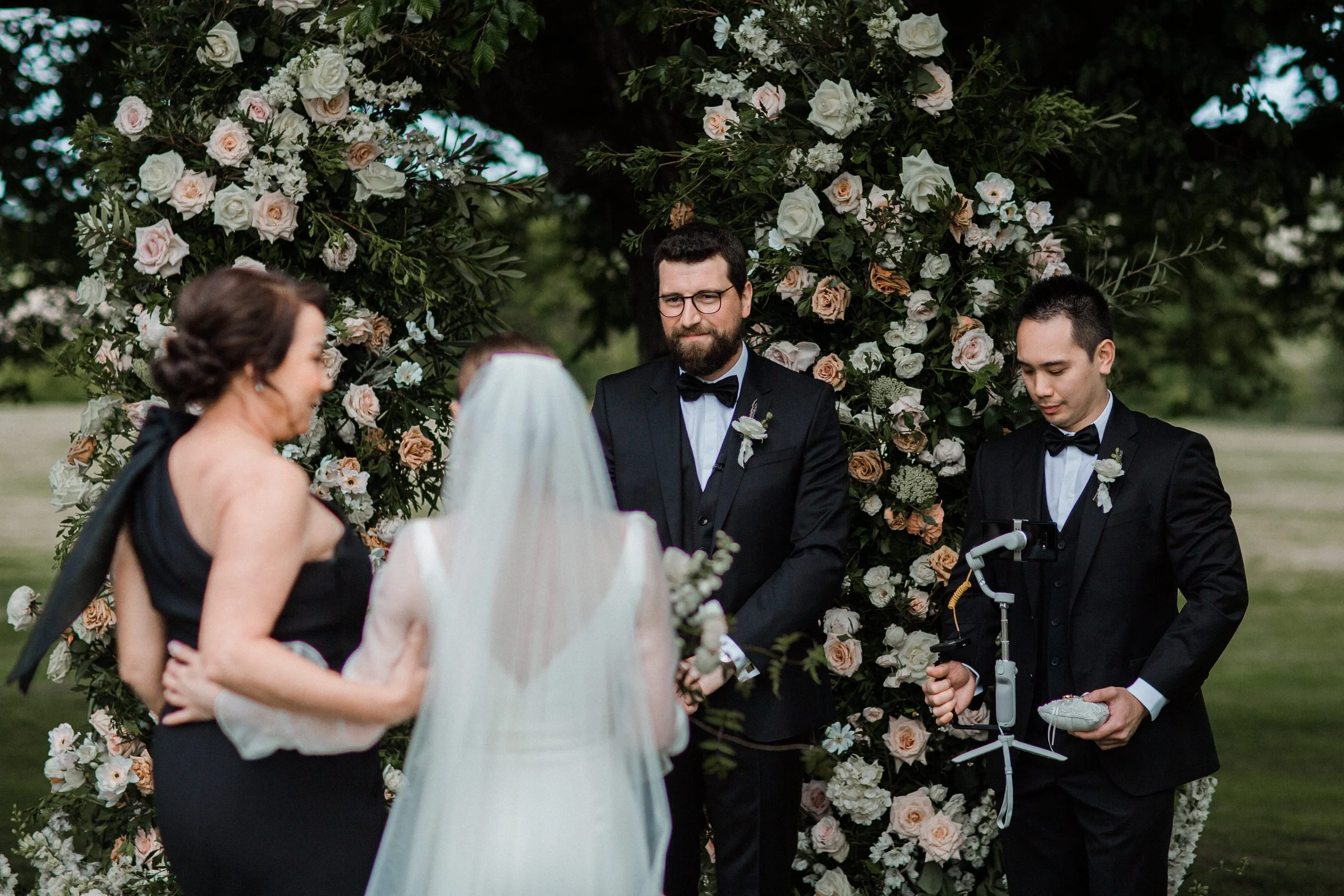 A wedding ceremony outdoors with a floral arch, a couple exchanging vows, a woman in a black dress, a groom in a tuxedo, and an officiant, with a videographer recording the event.