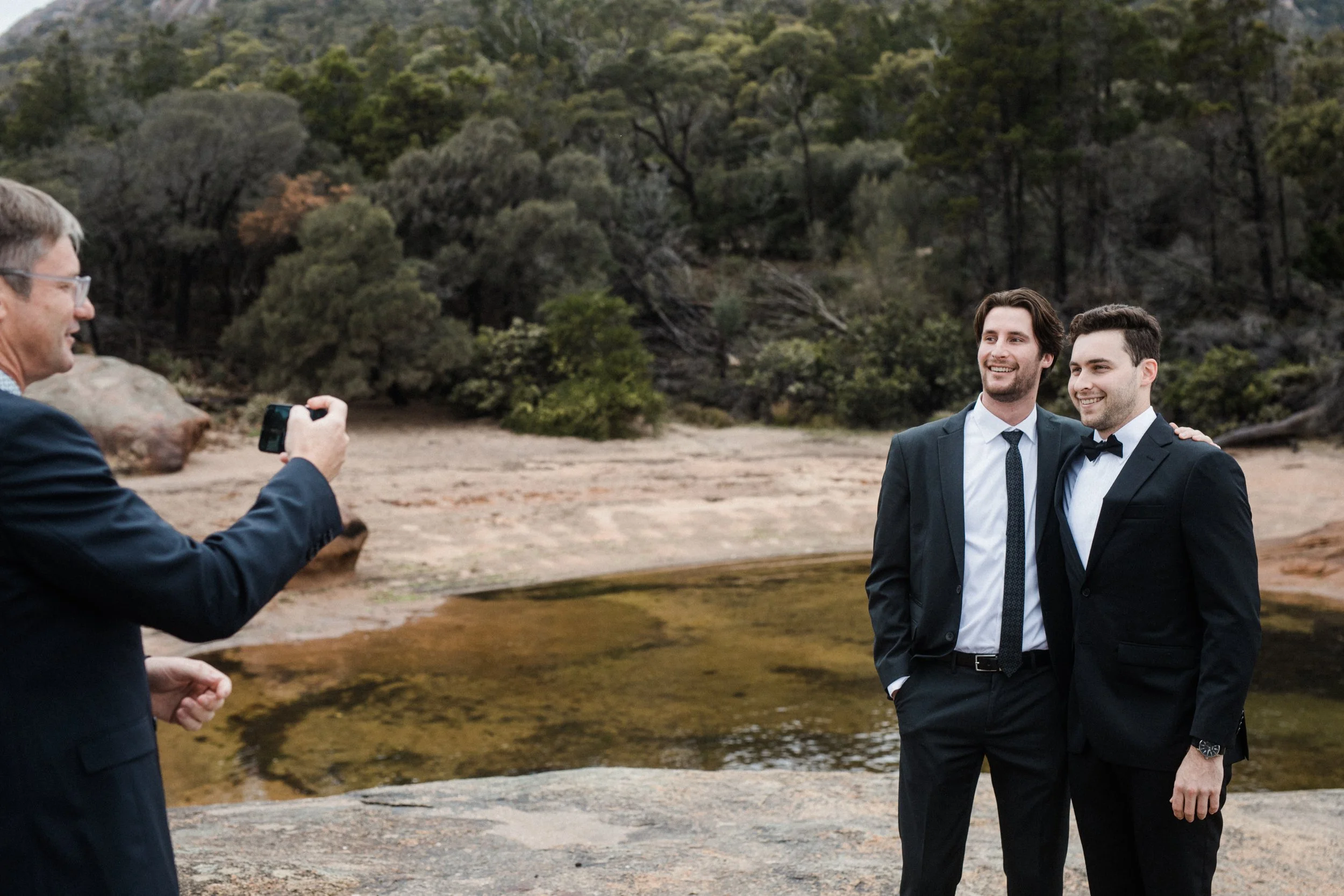 Two men in tuxedos with one wearing a bow tie and the other a regular tie, smiling and standing close together during a photo shoot in an outdoor natural setting, while a third man taking their photo with a smartphone.