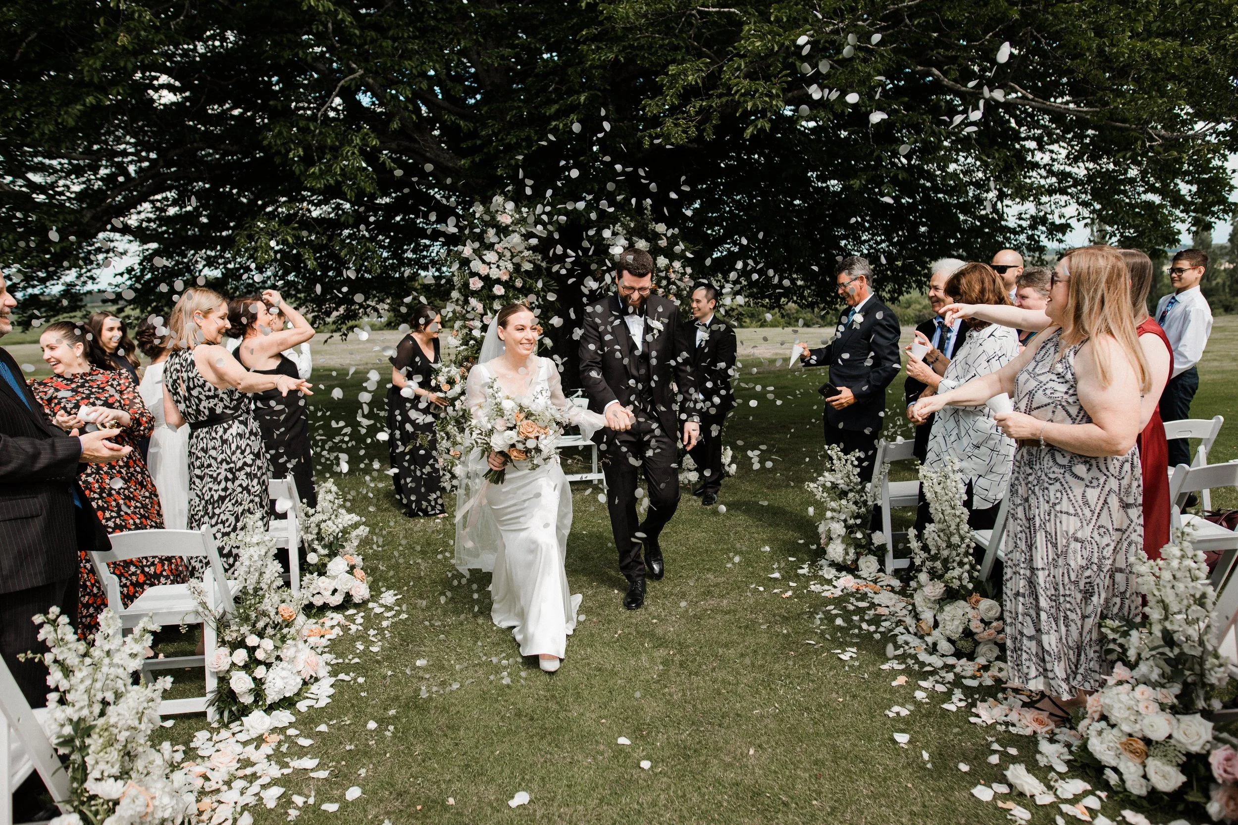 Bride and groom walking down the aisle after wedding ceremony surrounded by guests throwing flower petals under a large tree.
