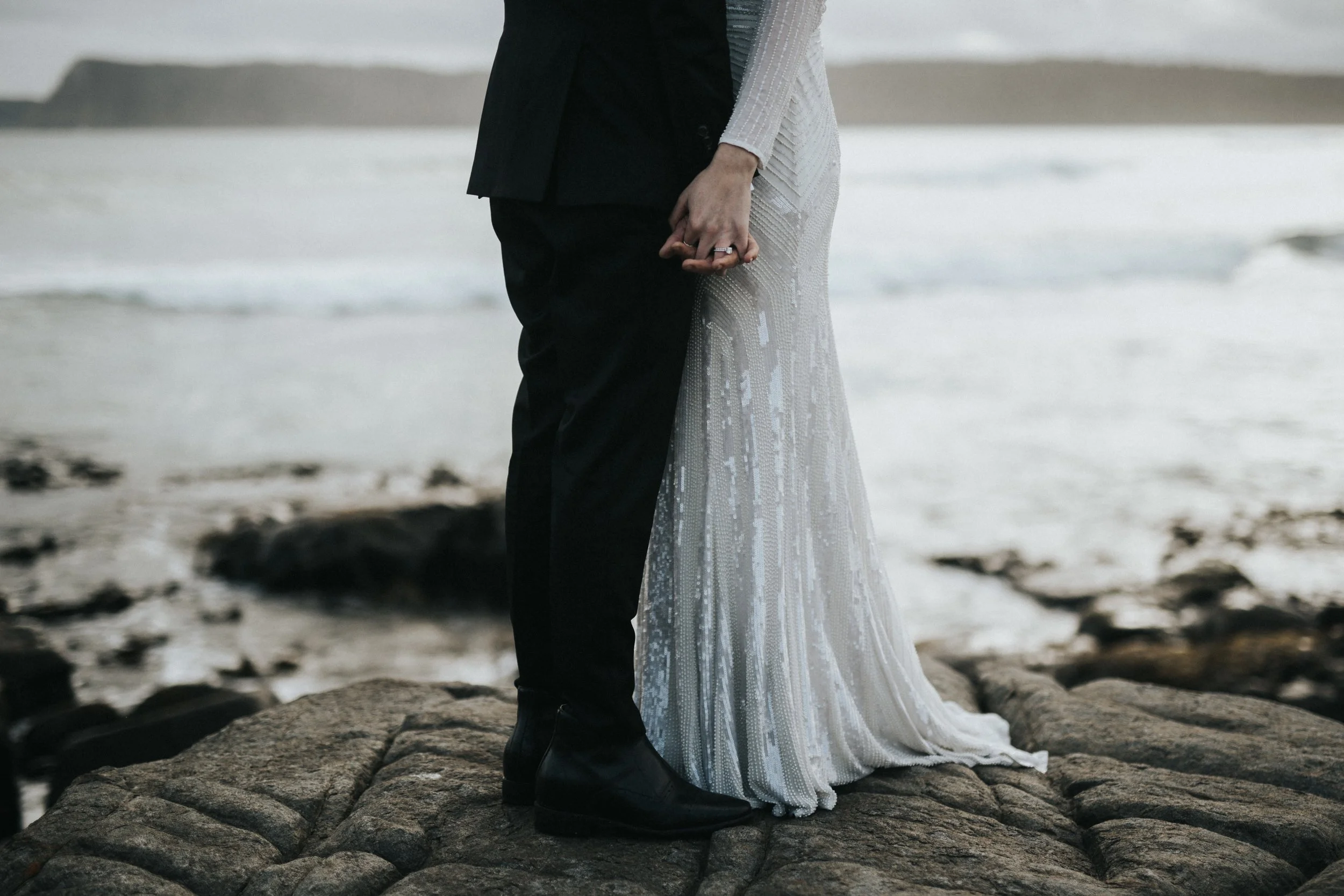 A couple dressed in wedding attire holding hands on a rocky beach, with the ocean and distant cliffs in the background.