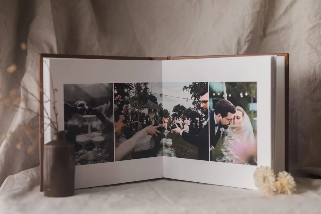 Open photo album with three wedding or engagement photos, surrounded by a small brown vase with flowers and some white flowers on a soft, light-colored fabric surface.