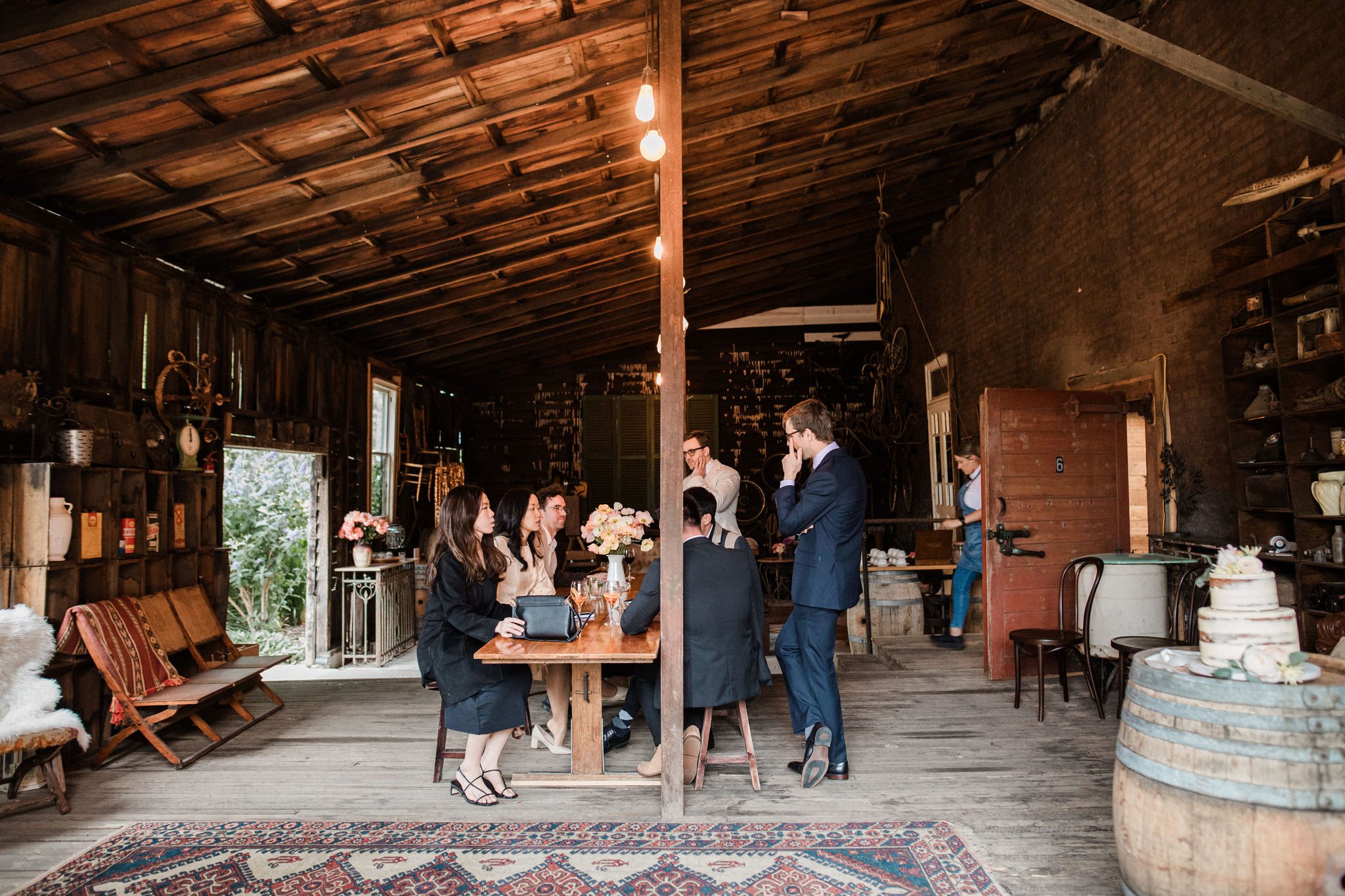 Group of people gathered around a wooden table in a rustic barn with wooden walls and ceiling, decorated with flowers and vintage items, having a meeting or celebration.