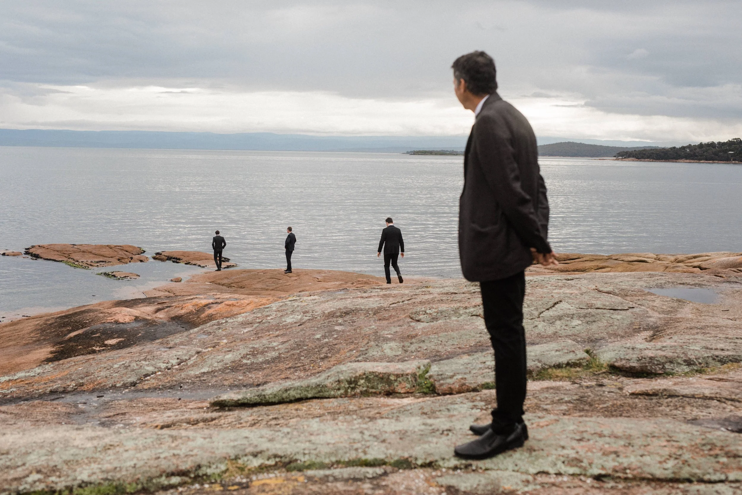 Four men in suits standing on rocky shoreline near water, with the sky overcast and distant land across the water.