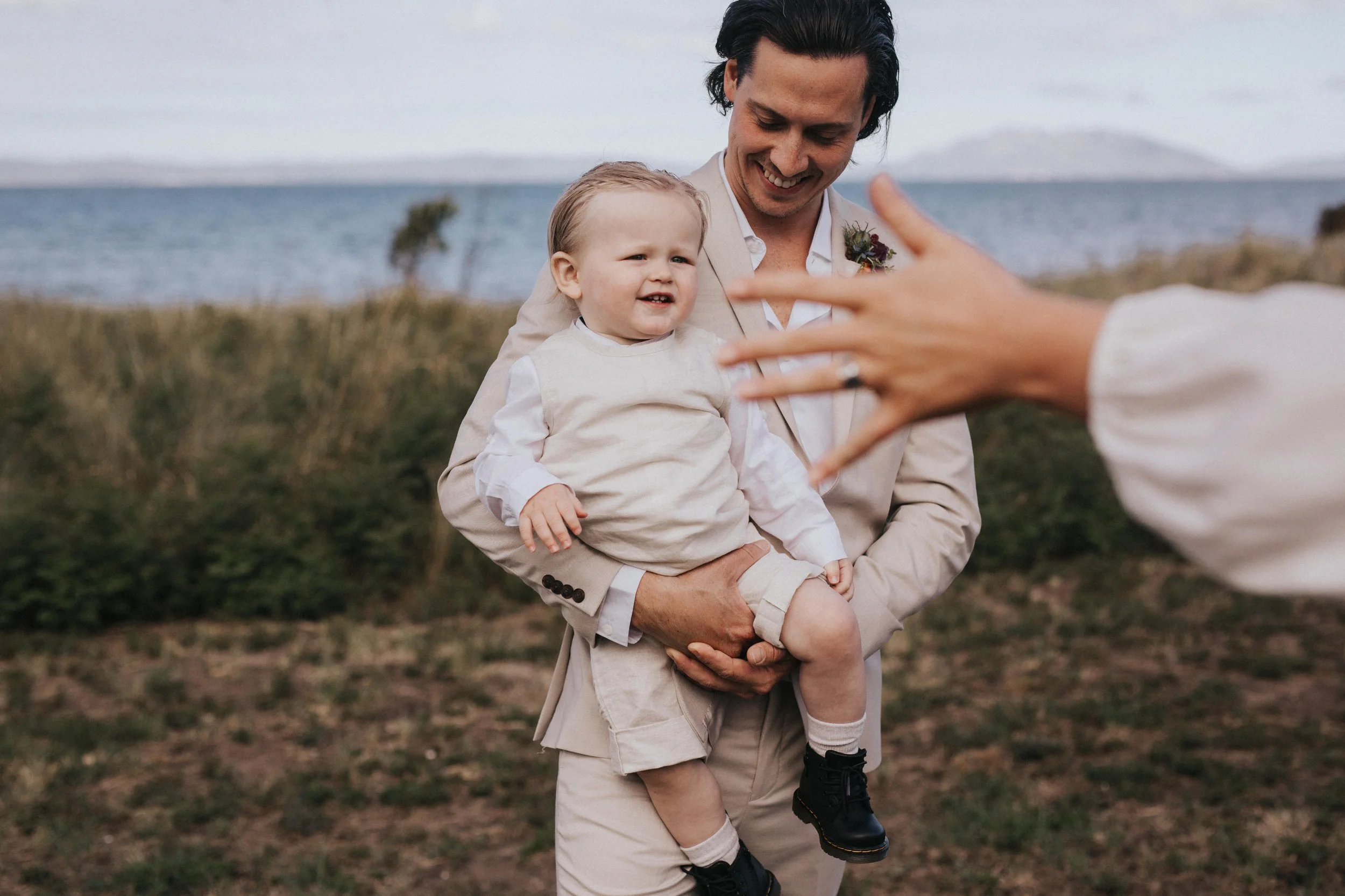 A man holding a young child outdoors near a body of water, with a blurred person reaching toward them in the foreground.