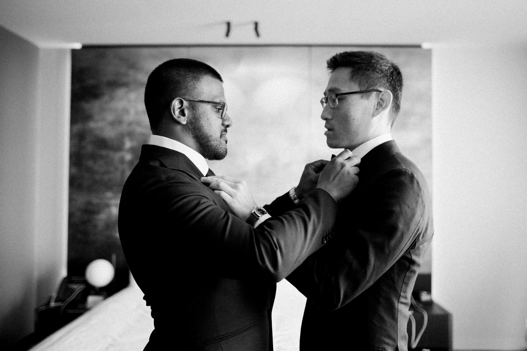 Two men in suits and glasses adjusting each other's ties in a hotel room.