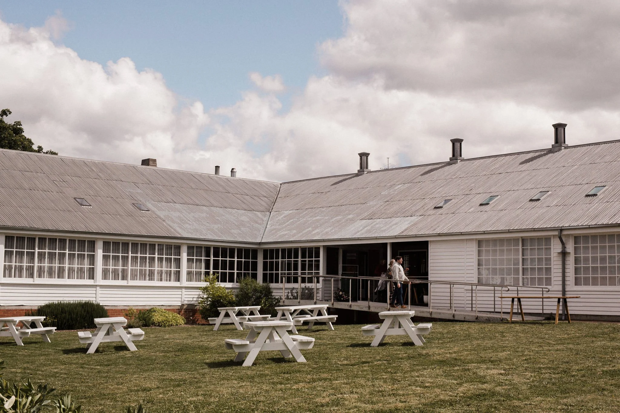 A large, white, single-story building with a aluminum roof and multiple skylights. In front, there are several white picnic tables on a grassy lawn, and a few people walking on a small deck.