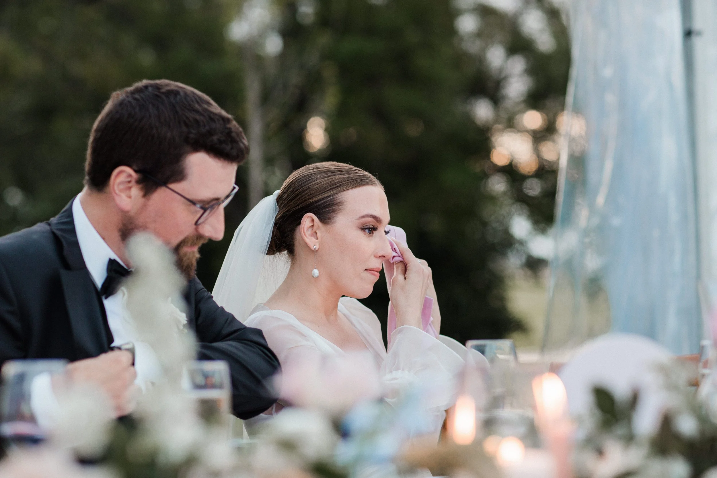 A bride with a white veil and pearl earrings, wiping her eye during a wedding ceremony, seated beside a groom in a black tuxedo, outdoors with blurred trees in the background.