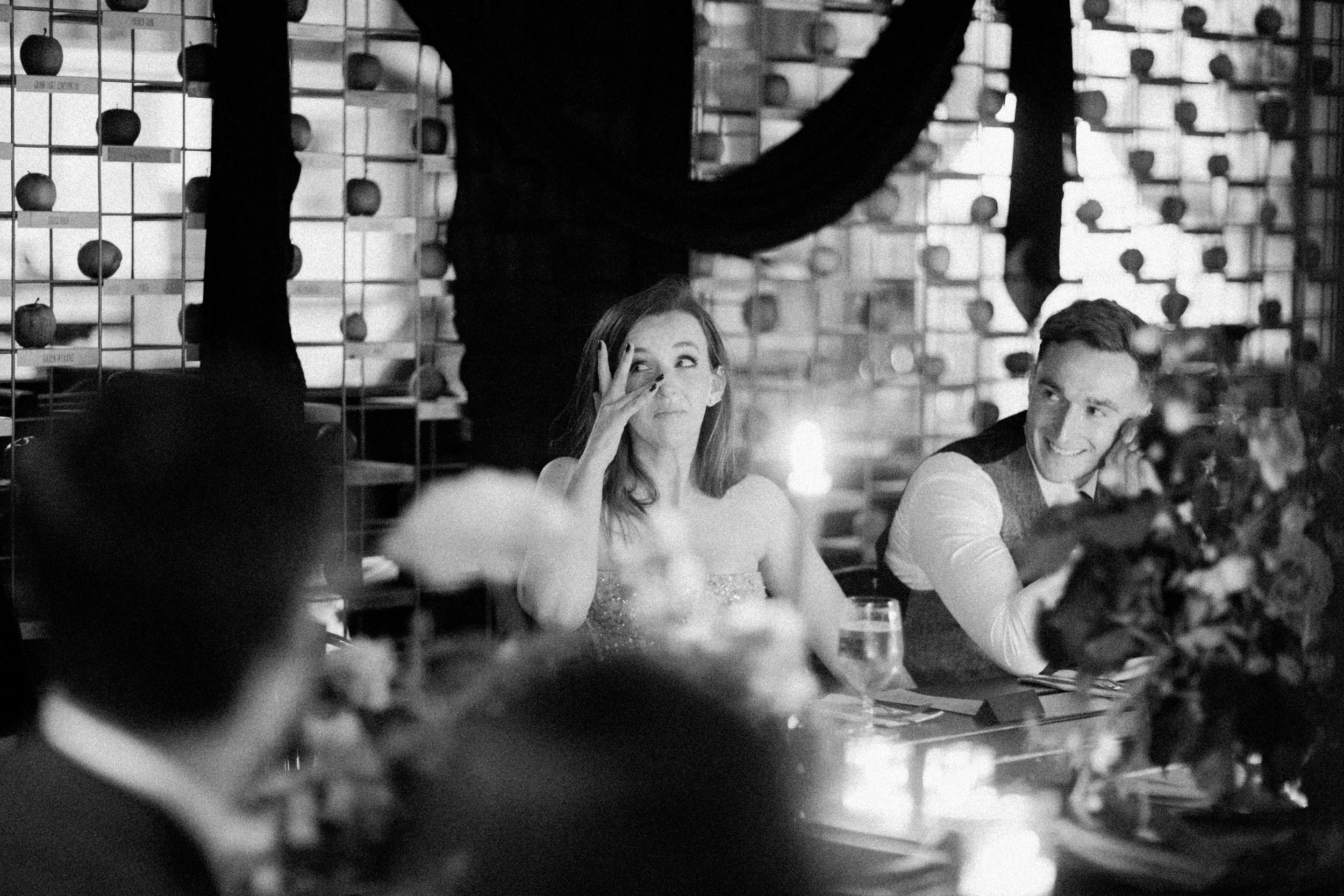 A black-and-white photo of people sitting at a dining table during a celebration or event, with a woman making a playful gesture near her eye and a man smiling nearby. In the background, there are decorative elements and shelves with objects.