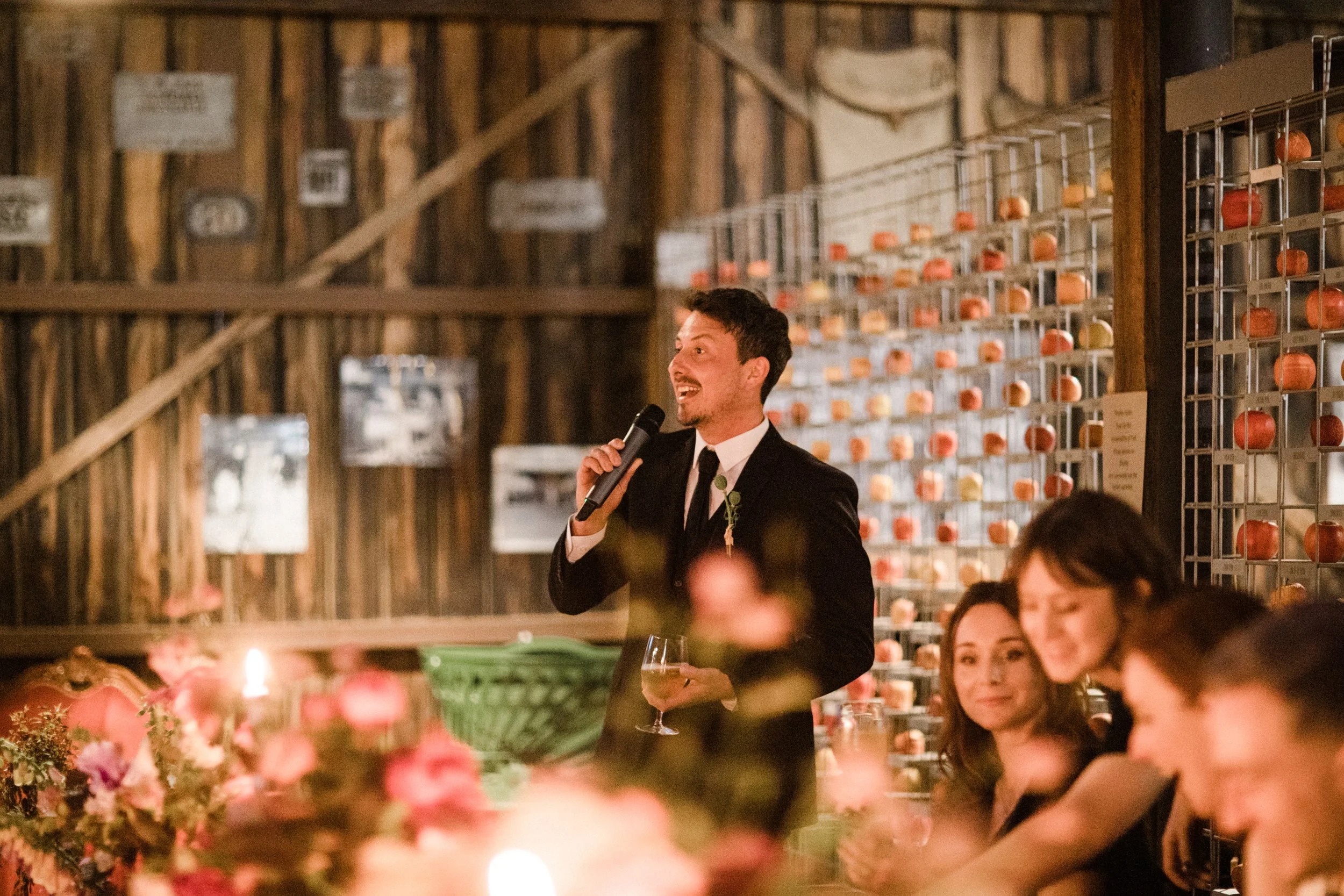 A man in a black suit with a tie, holding a glass of wine and speaking into a microphone at a wedding reception or celebration, with guests sitting at tables and a backdrop of wine bottles and wooden walls.