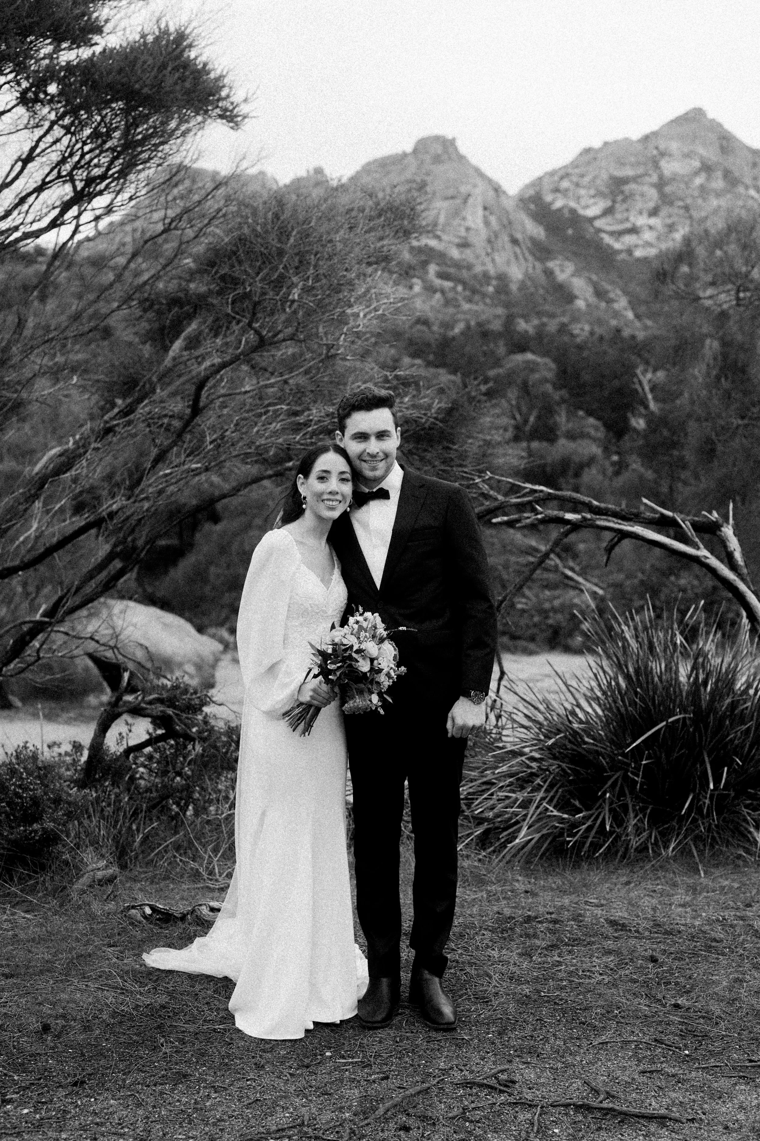 Black and white photo of a bride and groom standing outdoors in front of trees and mountains, smiling. The bride holds a bouquet of flowers, and the groom is dressed in a tuxedo.
