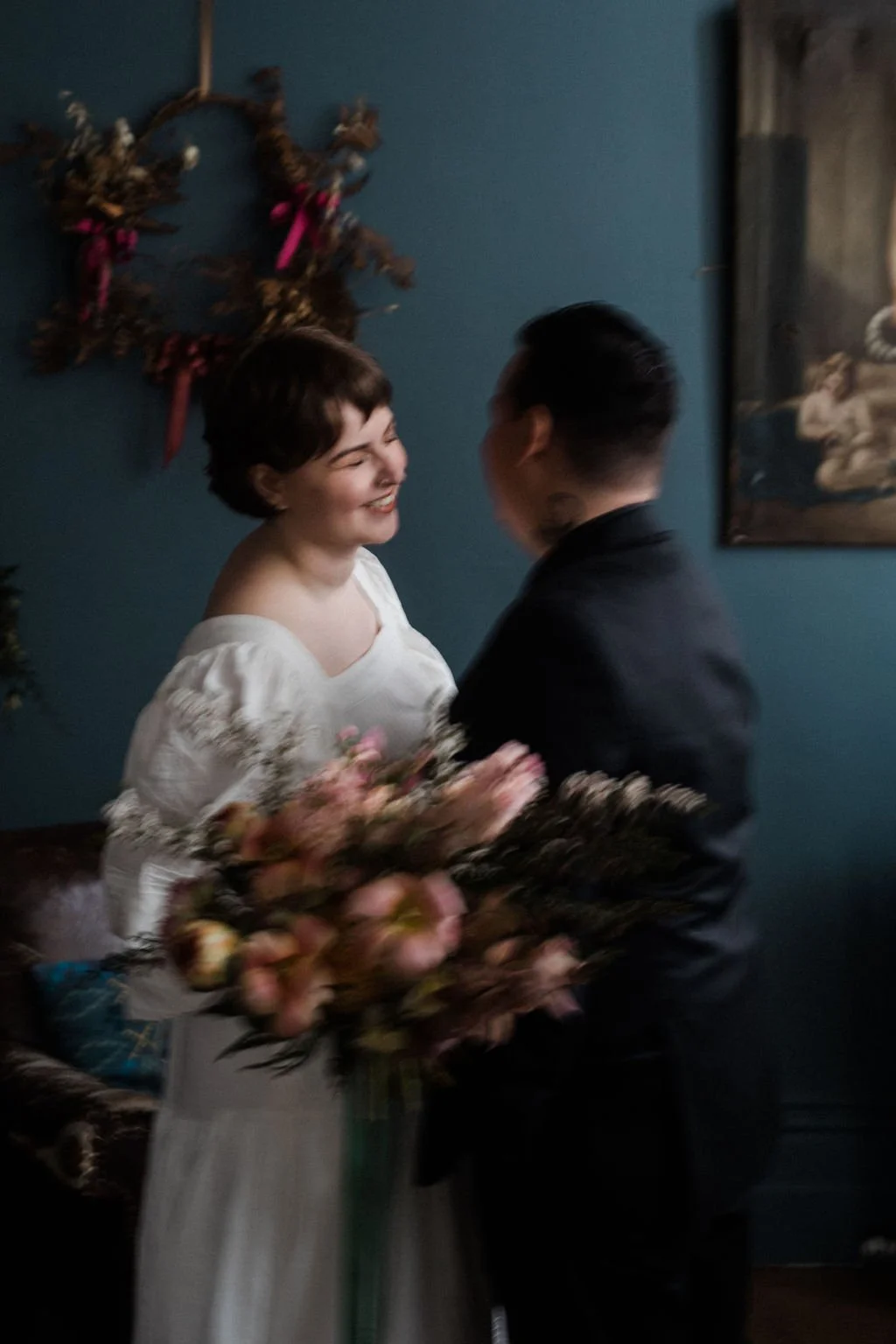 A woman in a white dress holding a bouquet of pink flowers, smiling at a man dressed in black, both standing indoors with blue walls and decorative wall hangings in the background.