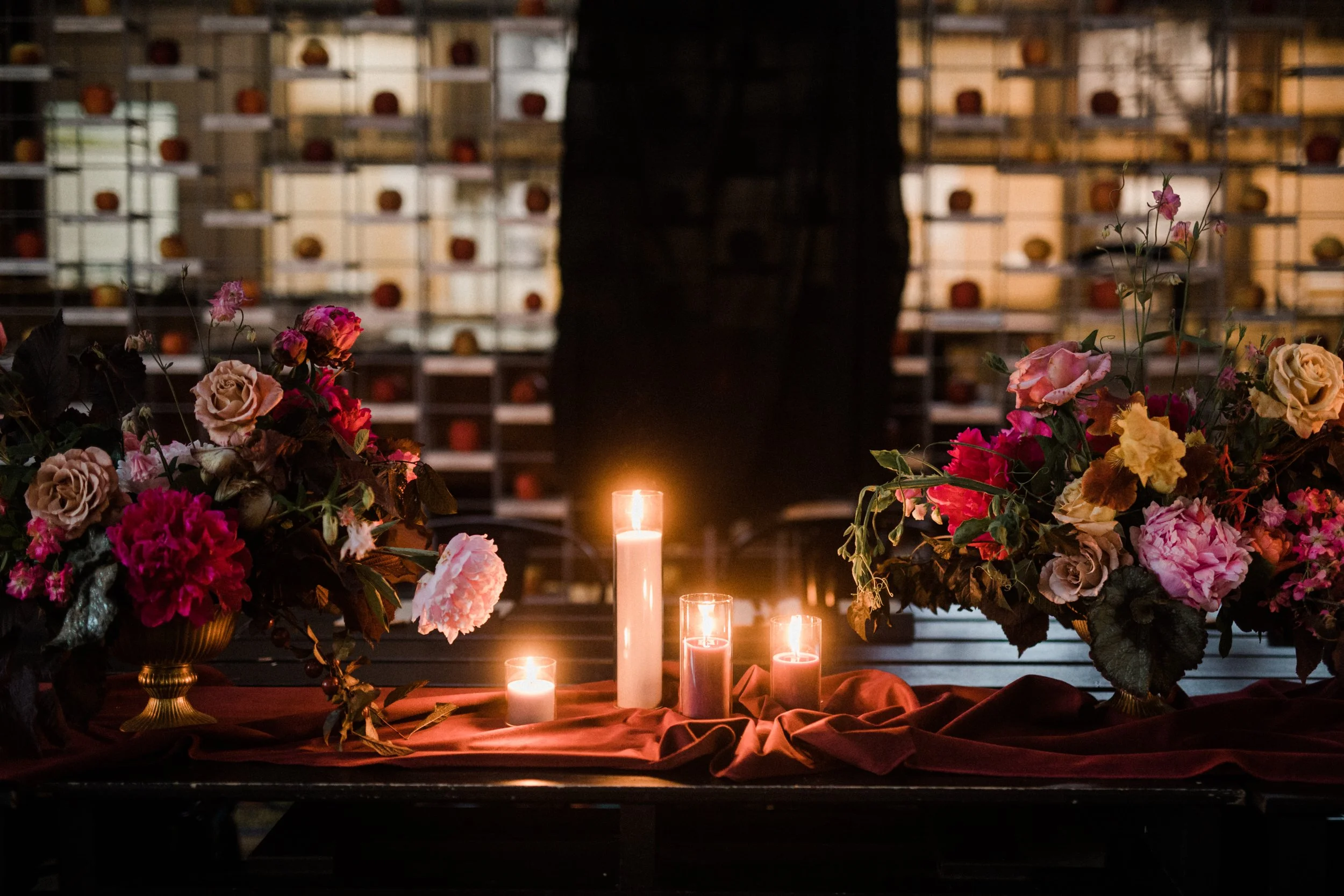 Flowers and lit candles on a table with a backdrop of shelves filled with pumpkins.