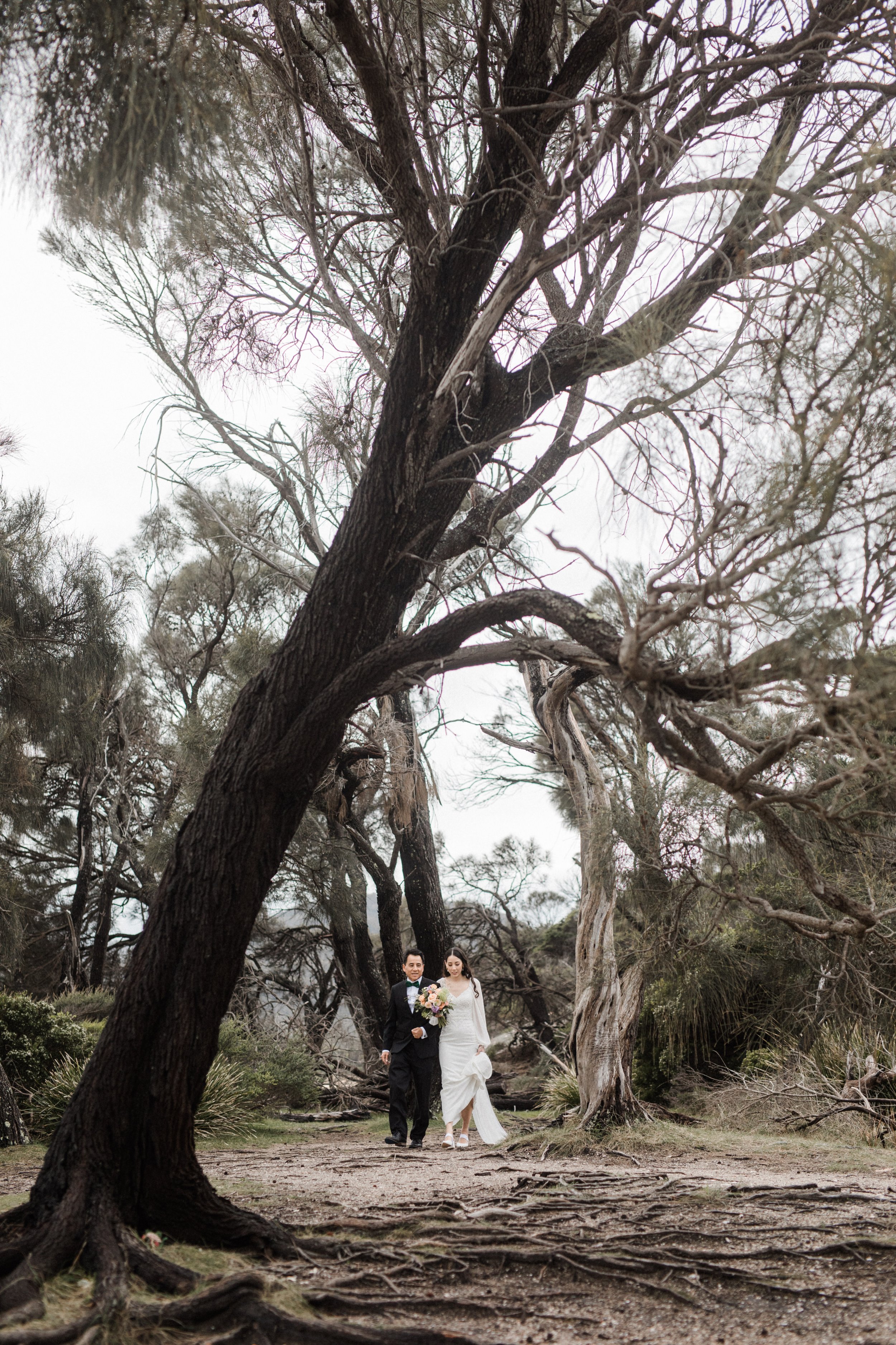 A bride and groom walking together outdoors on a dirt path surrounded by leafless trees, with the bride holding a bouquet of flowers.