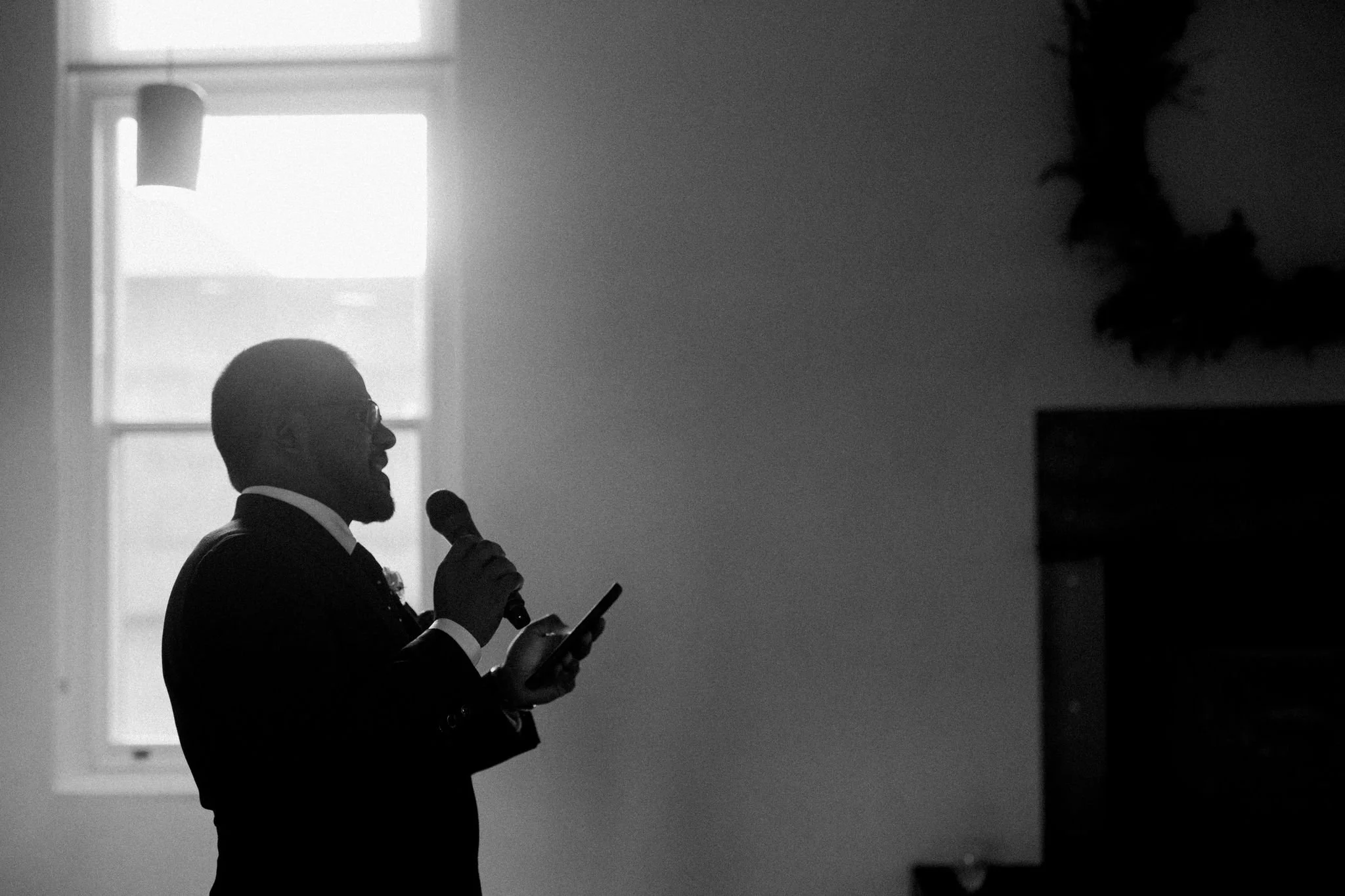 Silhouette of a man in a suit holding a microphone and a smartphone, speaking or giving a presentation in front of a window.