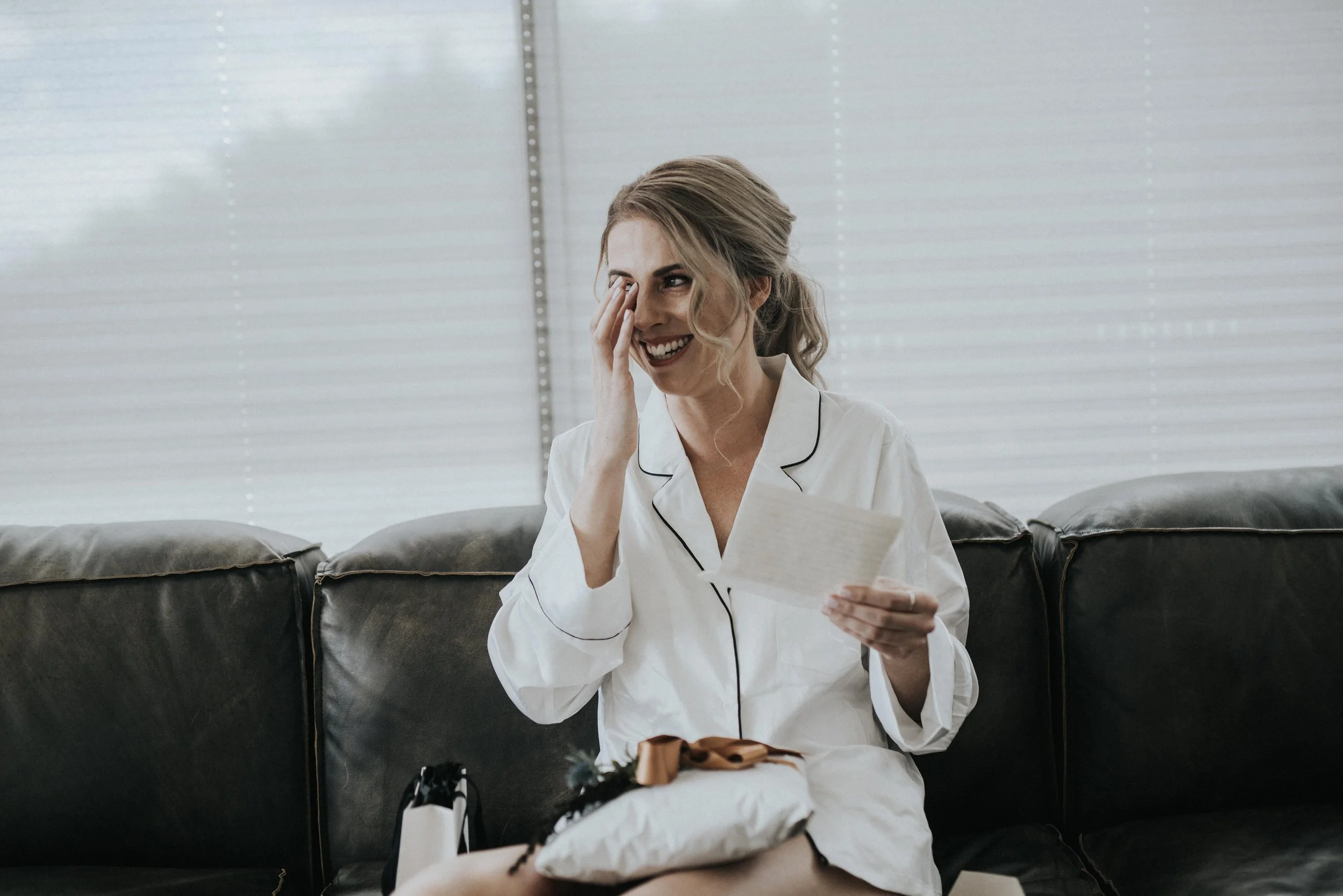 A woman in white pajamas sitting on a black leather couch, holding a piece of paper and smiling while wiping tears from her eye in a bright room with white blinds.