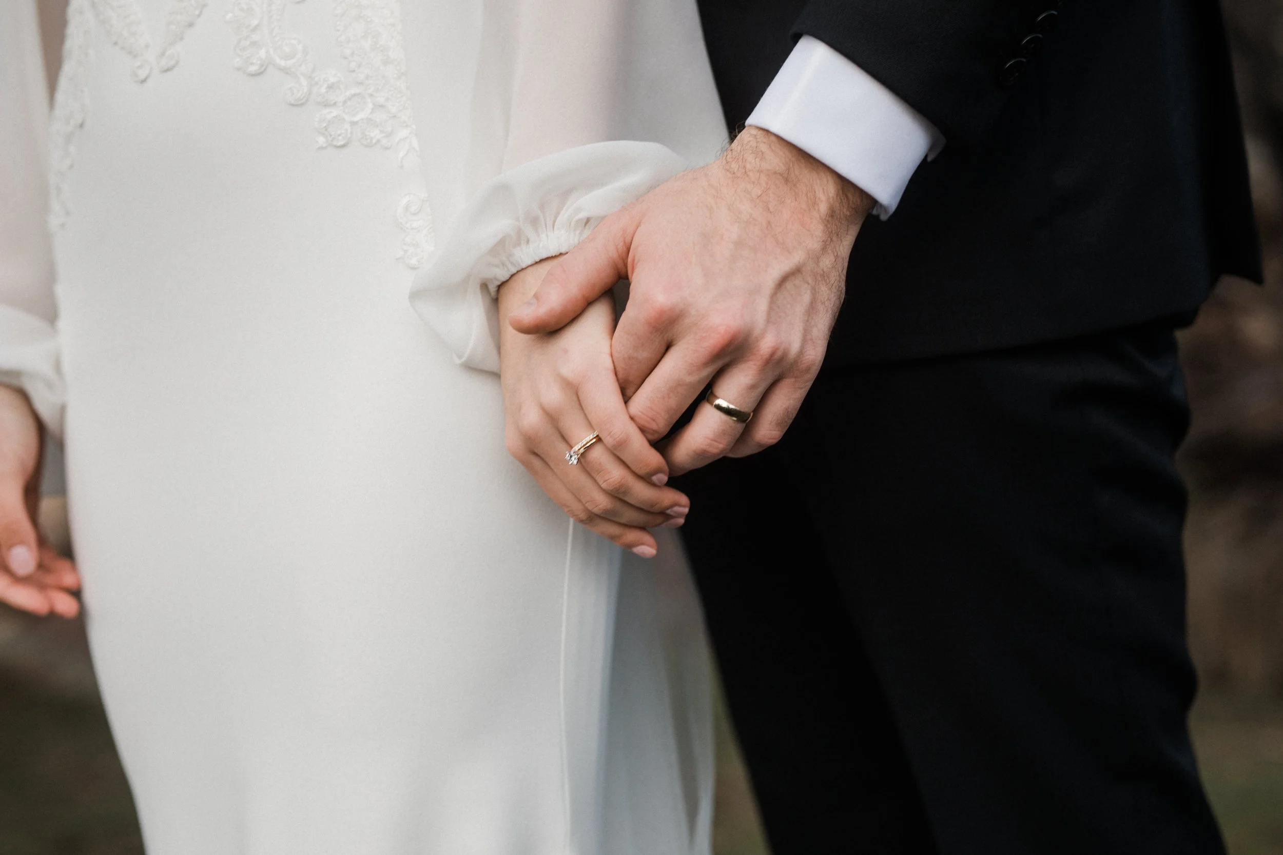 Close-up of a man and woman holding hands, showing wedding rings, with the bride in a white dress and the groom in a black suit.
