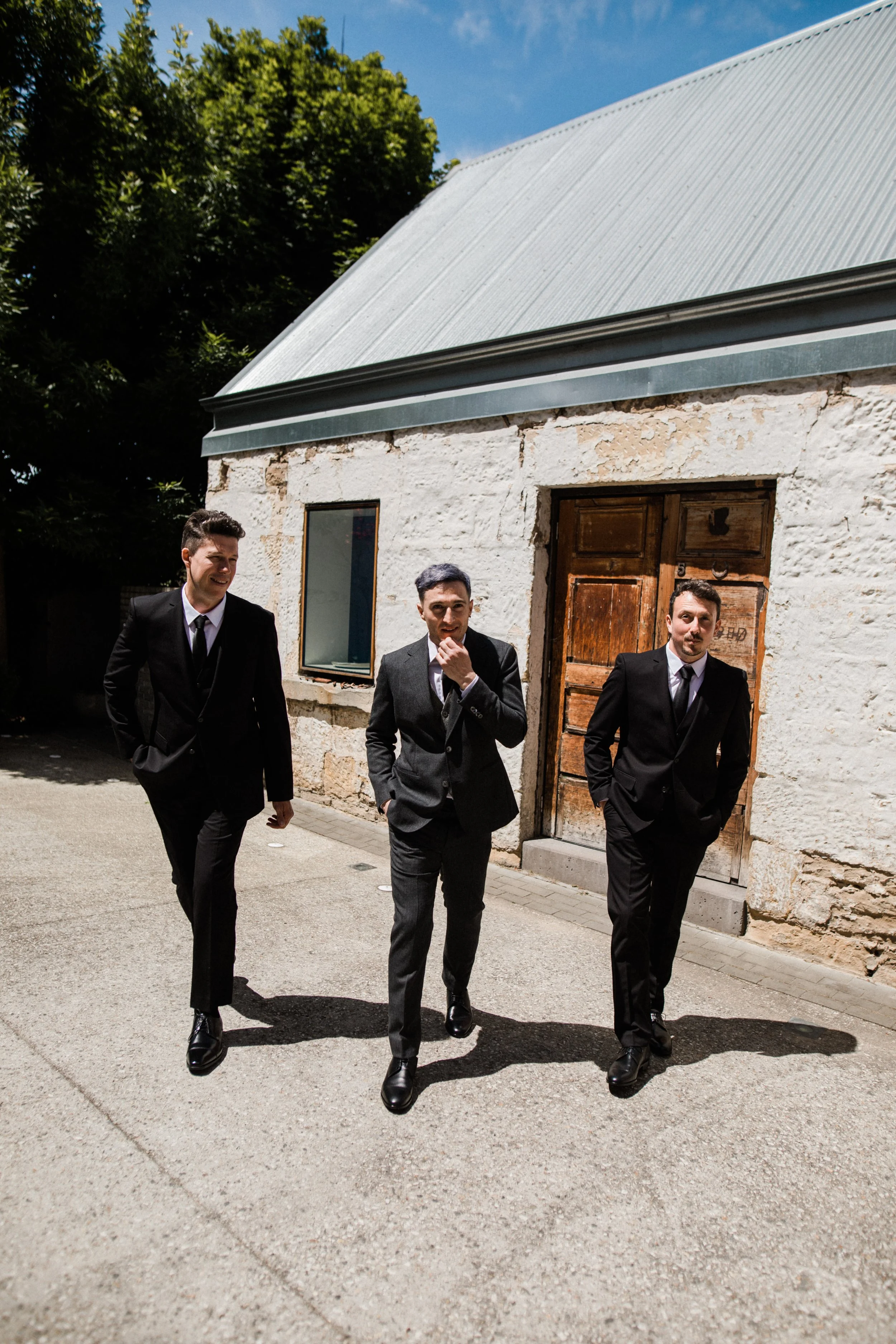 Three men in black suits walking outside on a sunny day, in front of a rustic white stone building with a metal roof and wooden door.