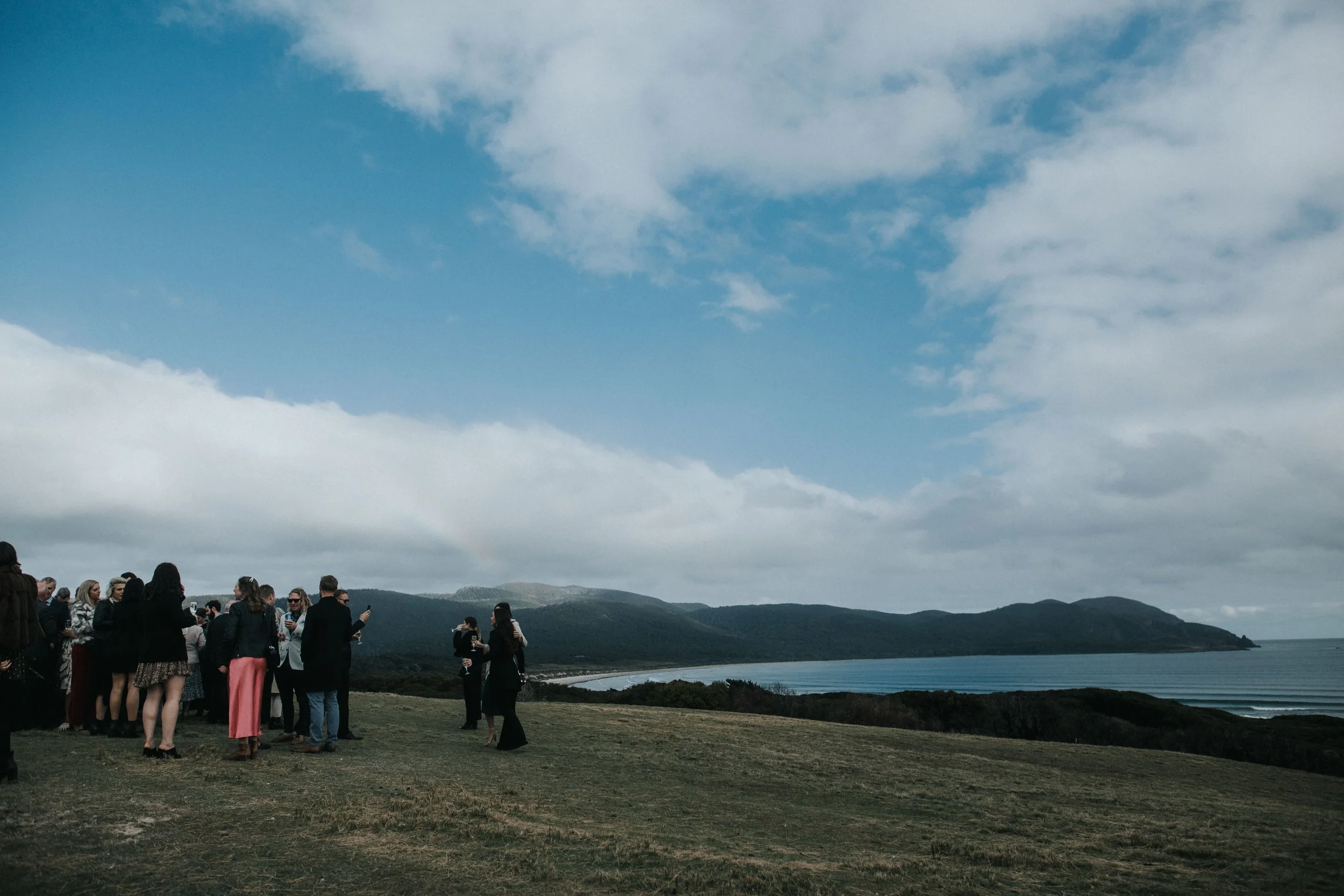 Group of people gathered outdoors on a grassy hill, with a view of hills, ocean, and partly cloudy sky in the background.