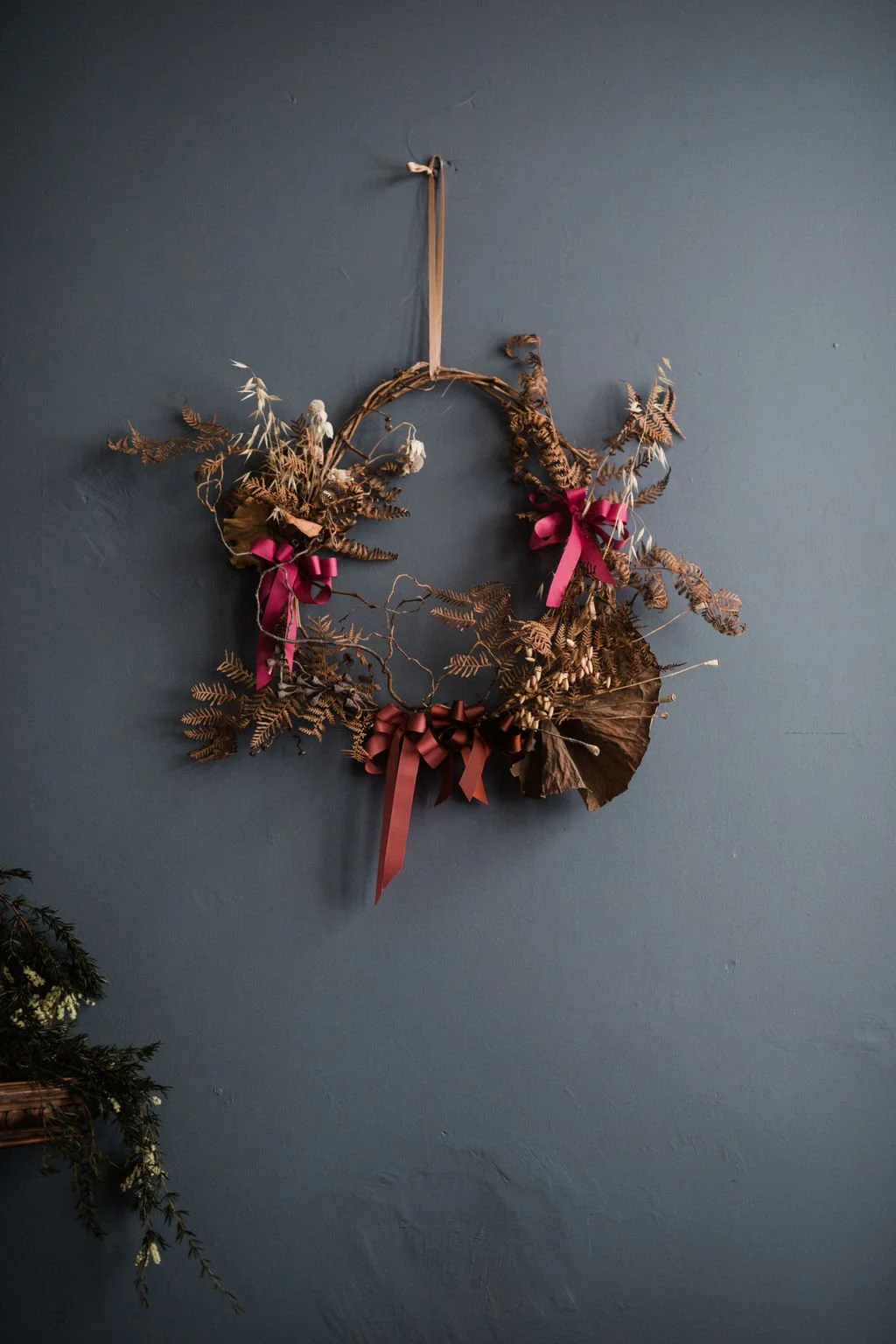 A decorative wreath made of brown dried leaves and ferns, tied with pink and brown ribbons, hanging on a dark blue wall.