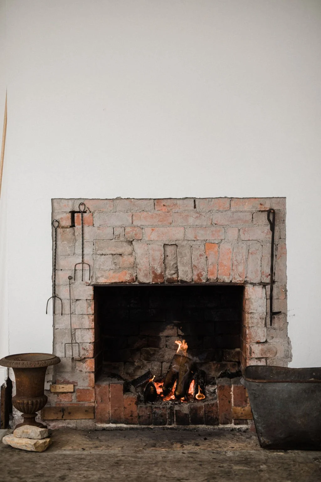 Brick fireplace with a small fire burning inside, white wall behind, and decorative items like a small ornamental pedestal and a large metal container on either side.
