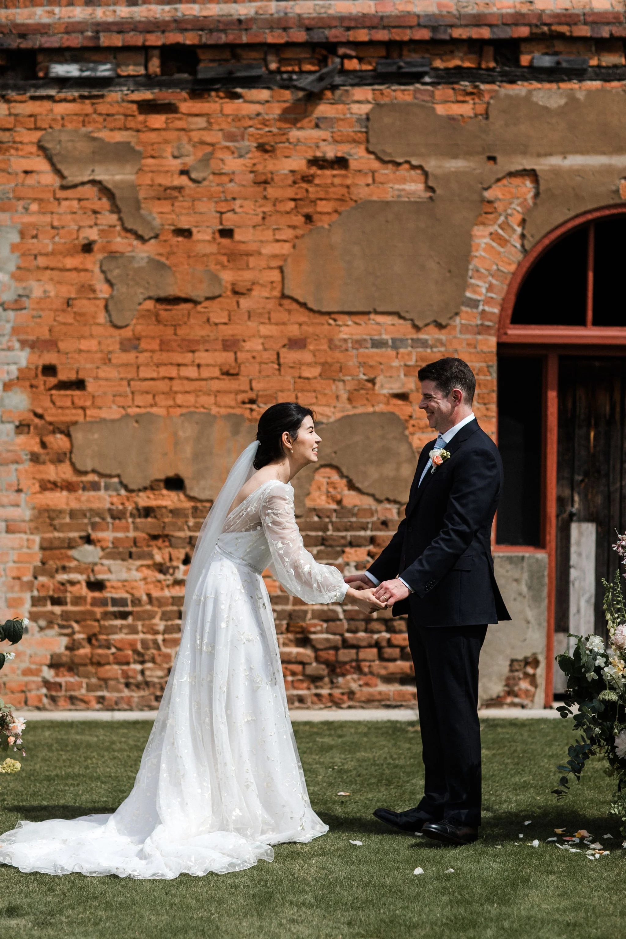 A bride and groom holding hands and smiling at each other during their wedding ceremony outdoors with a brick wall in the background.
