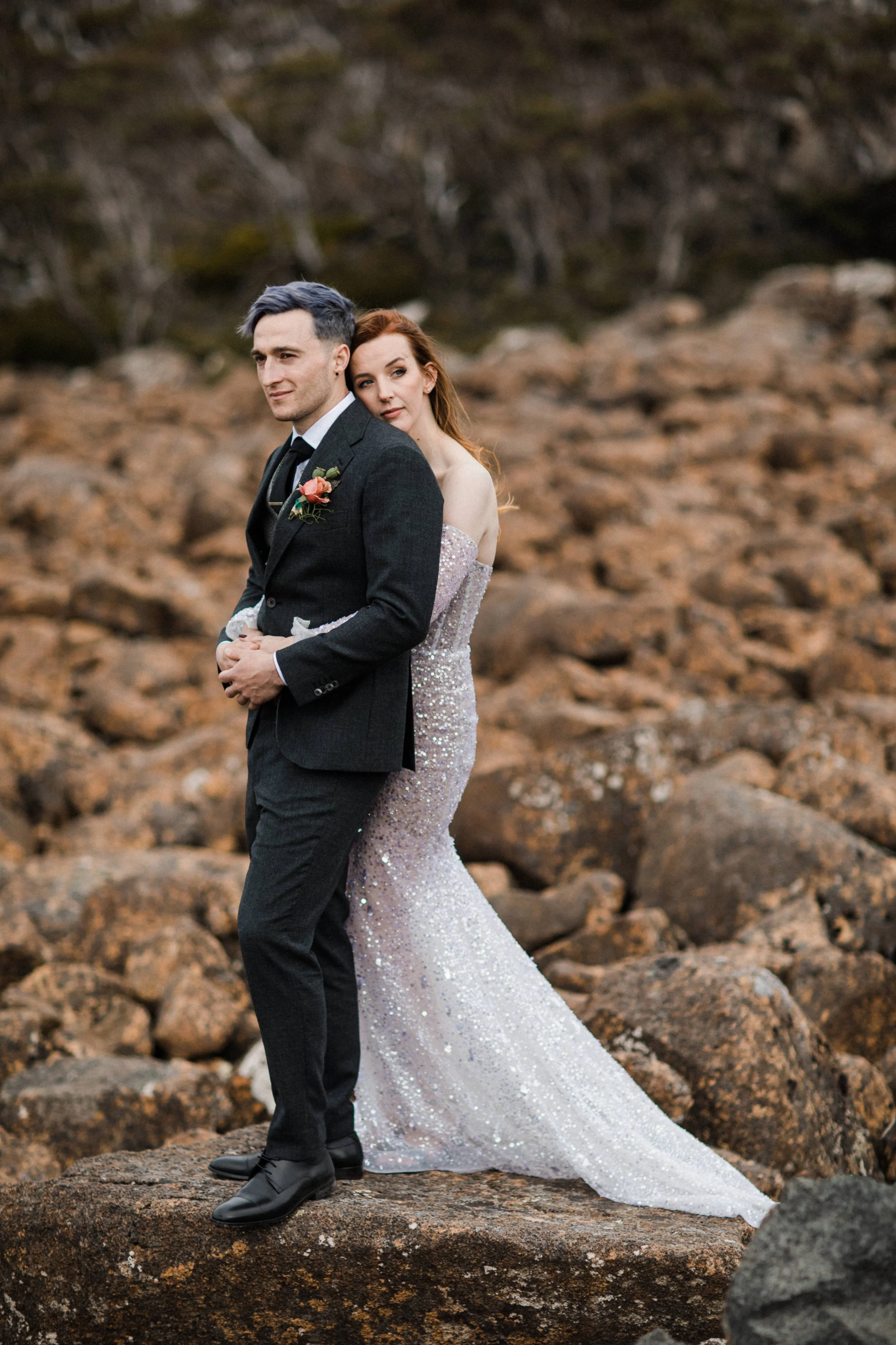 A bride and groom standing on rocks outdoors, the groom in a black suit with boutonniere, and the bride in a sparkly strapless wedding gown with train, embracing amid a rocky landscape.