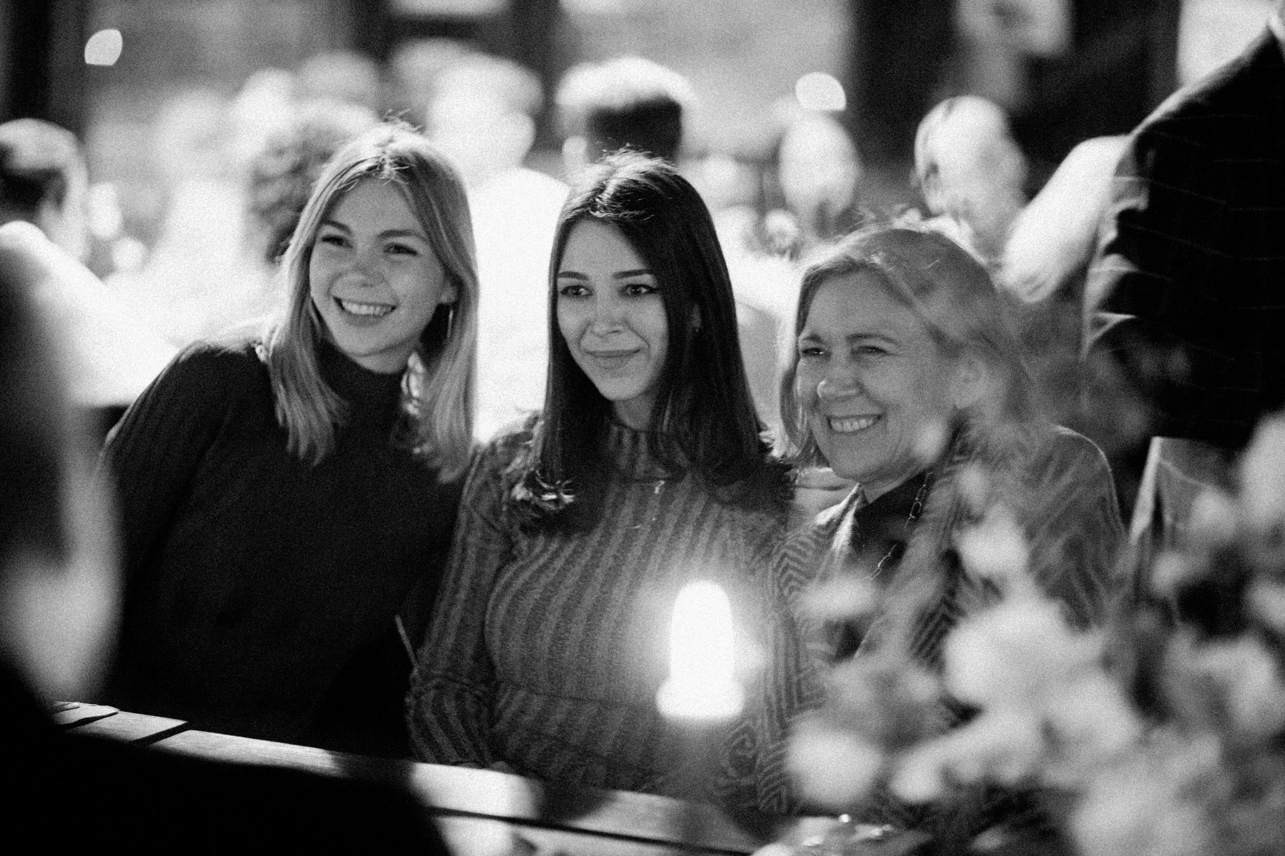 Three women smiling and enjoying a moment together at a social gathering, with blurred background and a candle on the table.