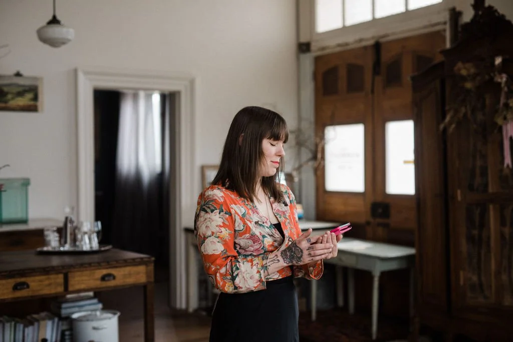 A woman with dark brown hair and bangs, wearing a floral patterned orange blazer, is looking at her pink smartphone in a cozy, rustic room with wooden furniture and windows.