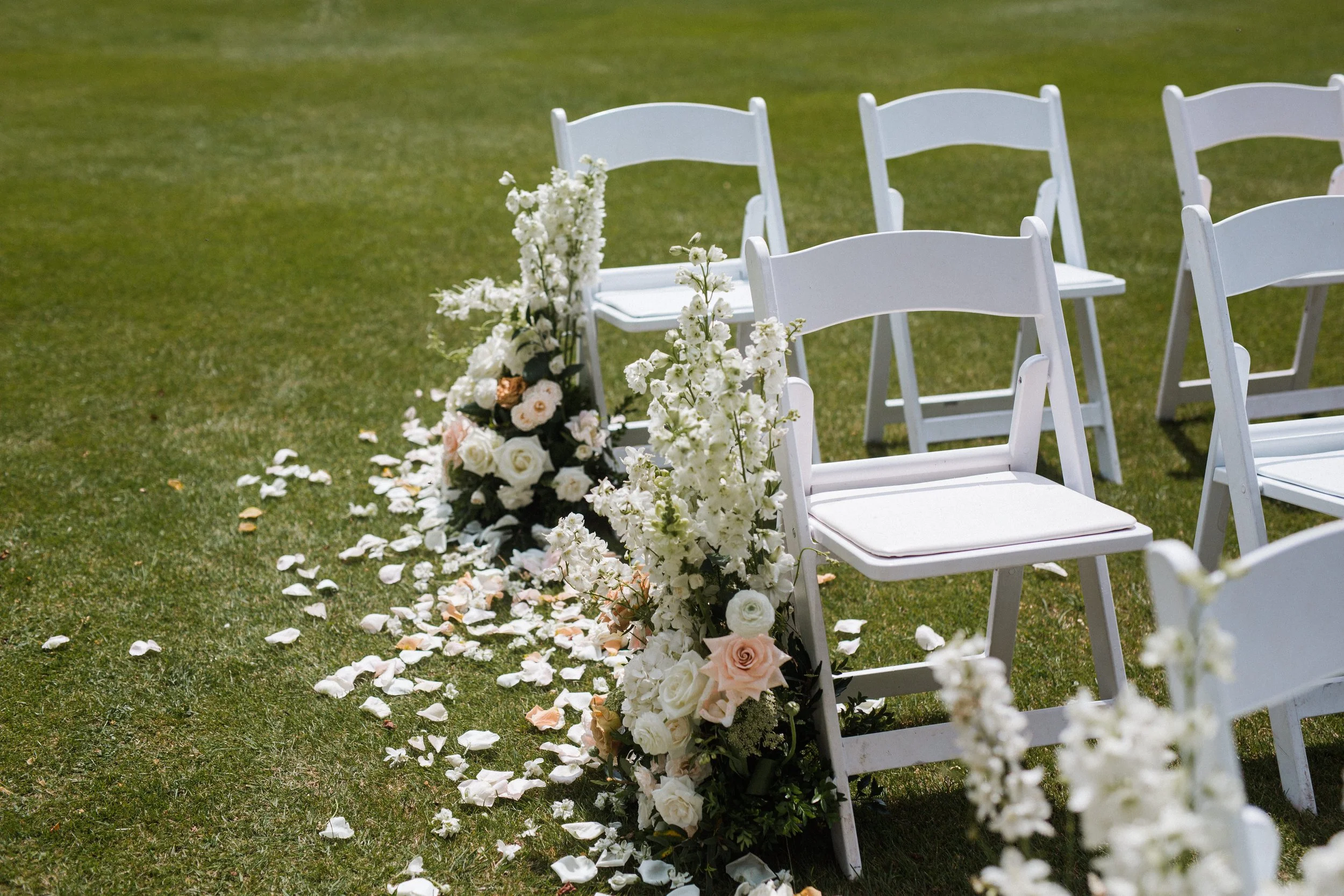 White folding chairs arranged outdoors with floral decorations and scattered flower petals on grass.